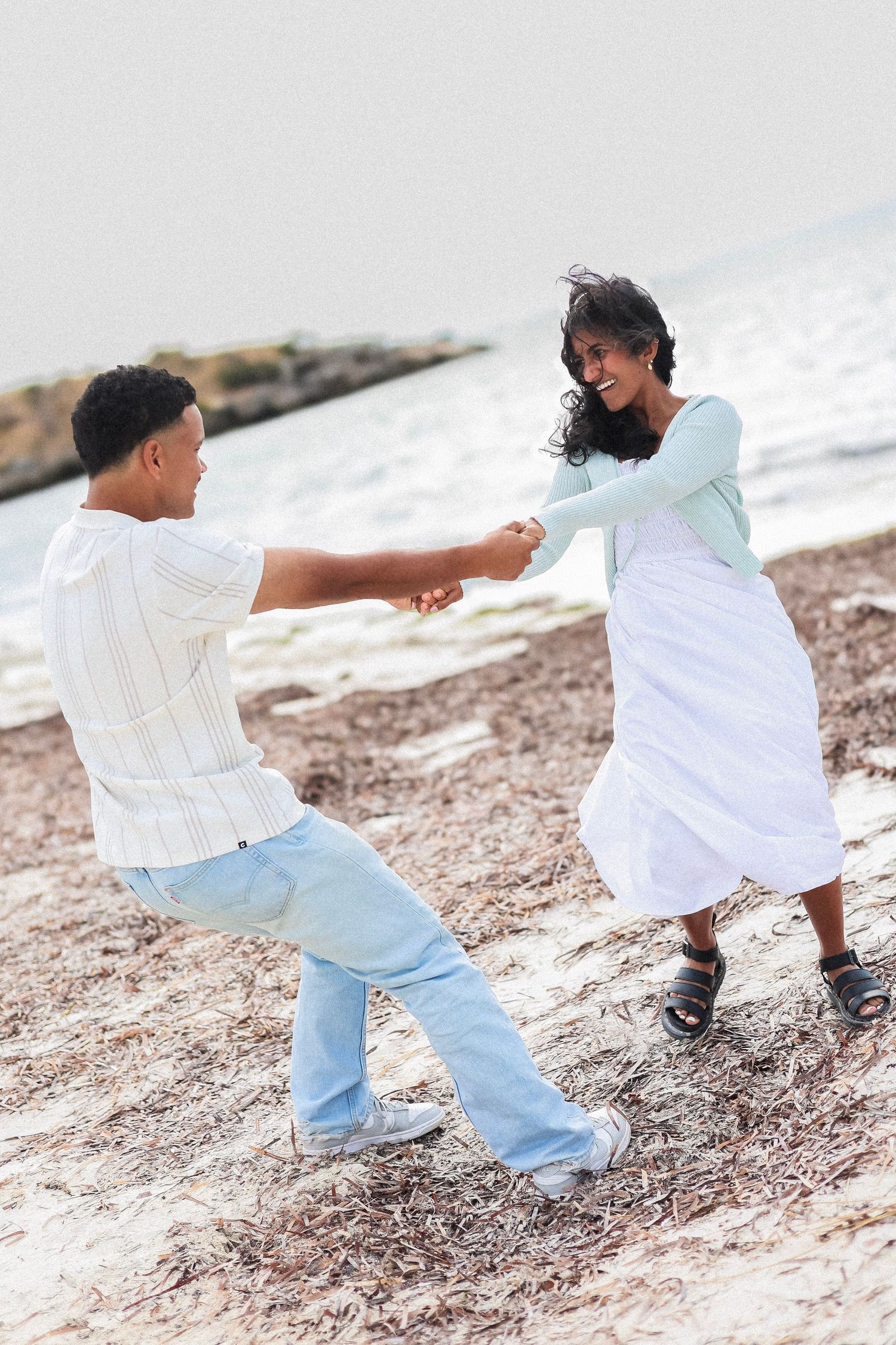 A joyful couple playfully dances and spins together on a rocky beach shoreline by the ocean.