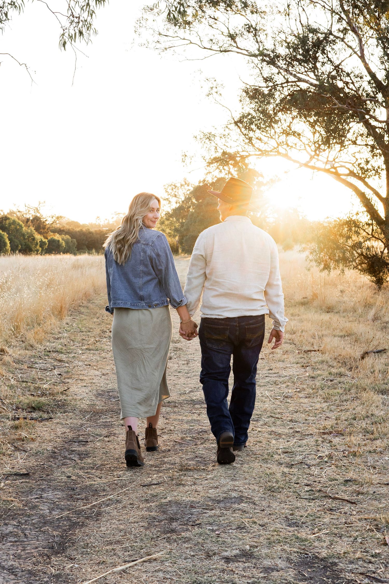 Couple walking hand in hand down a dirt path through golden fields toward a glowing sunset.