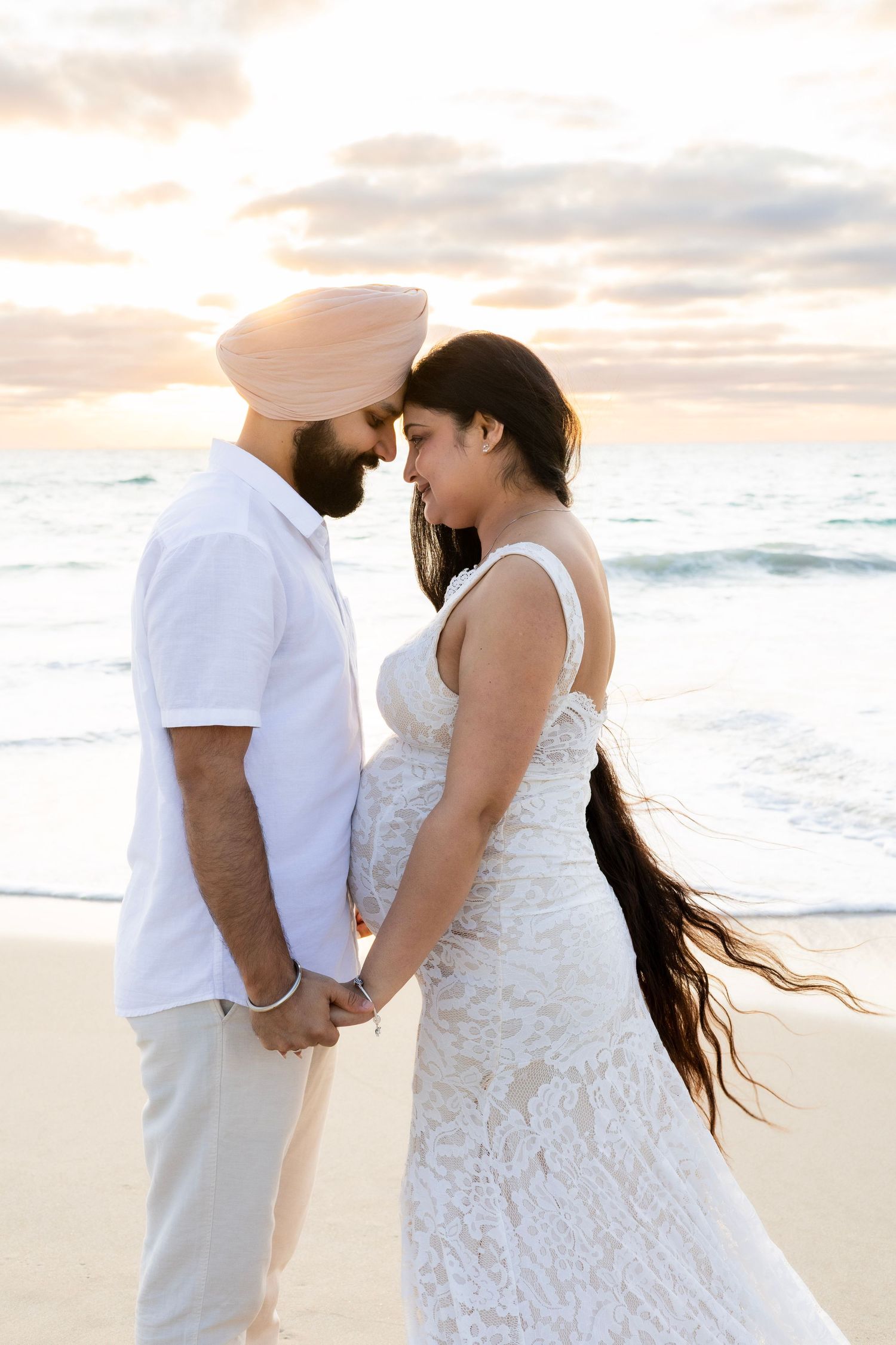 Expecting couple in white outfits holding hands facing each other on beach at golden sunset.