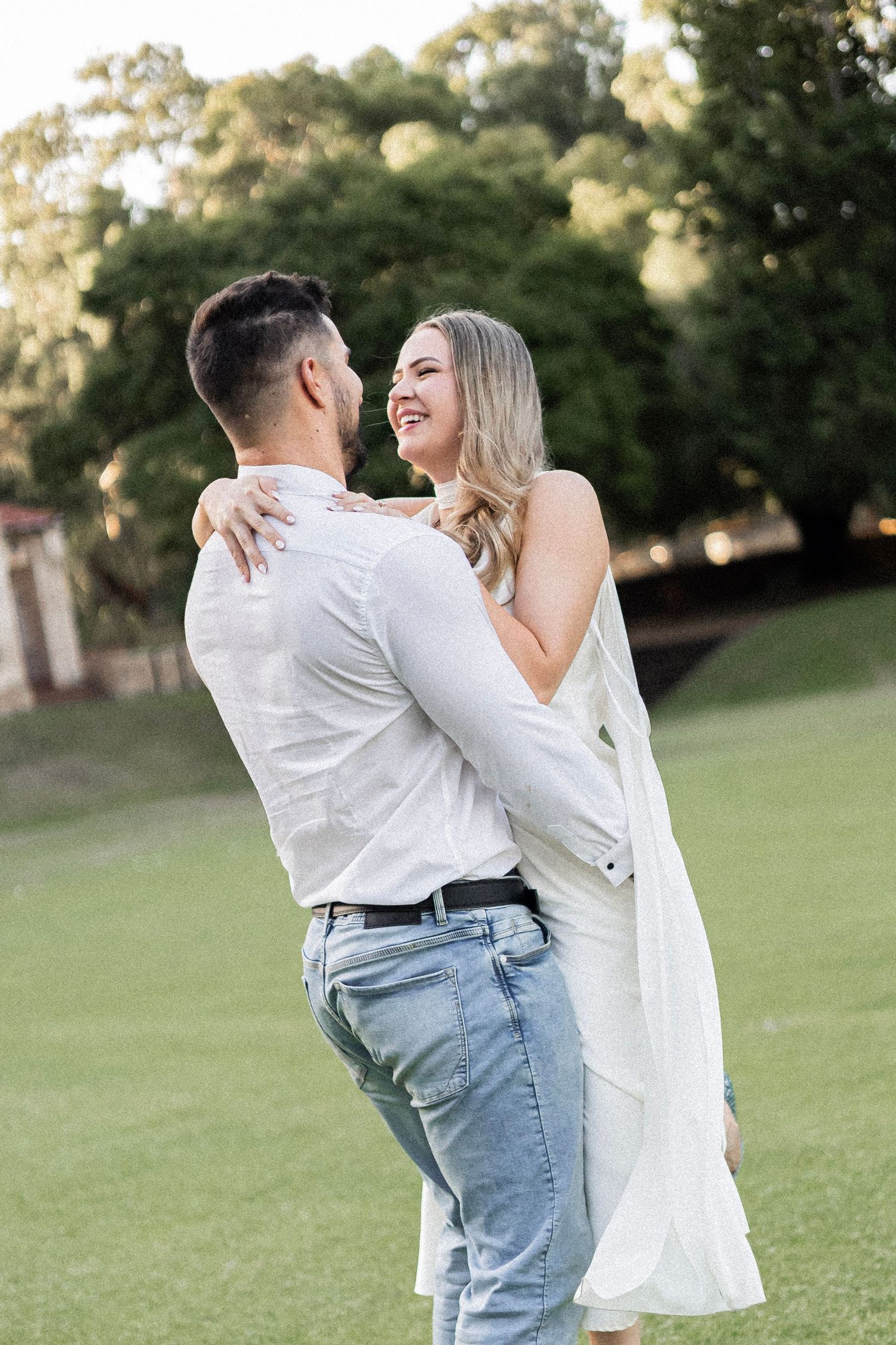 Joyful couple embracing and laughing together on a sunlit green lawn with trees behind them.
