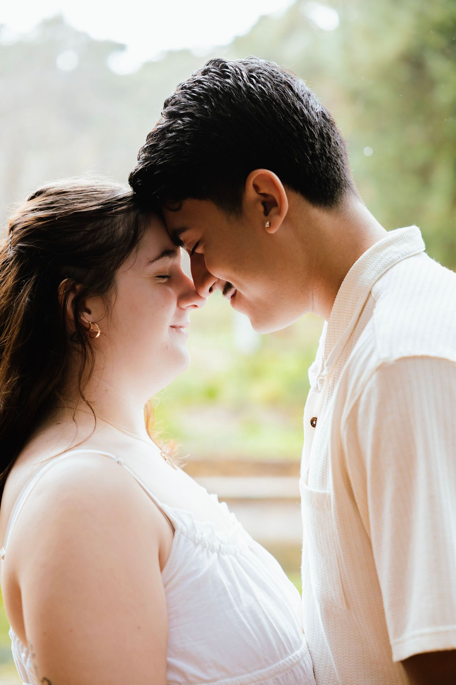 Young couple sharing a tender forehead-to-forehead moment outdoors, wearing white outfits in soft natural light.