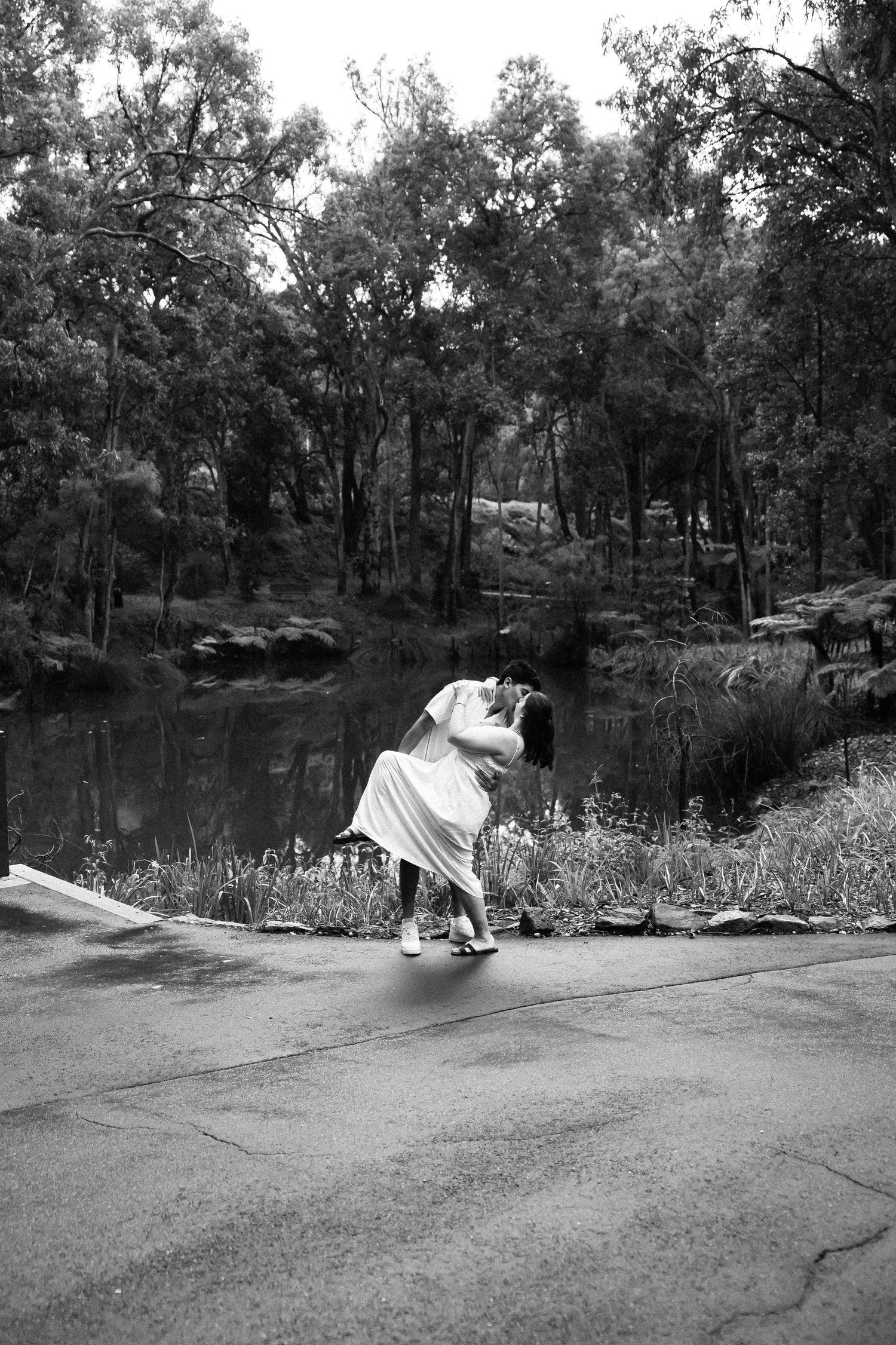 Black and white photo of a couple sharing a romantic dip kiss by a tranquil forest pond on their wedding day.