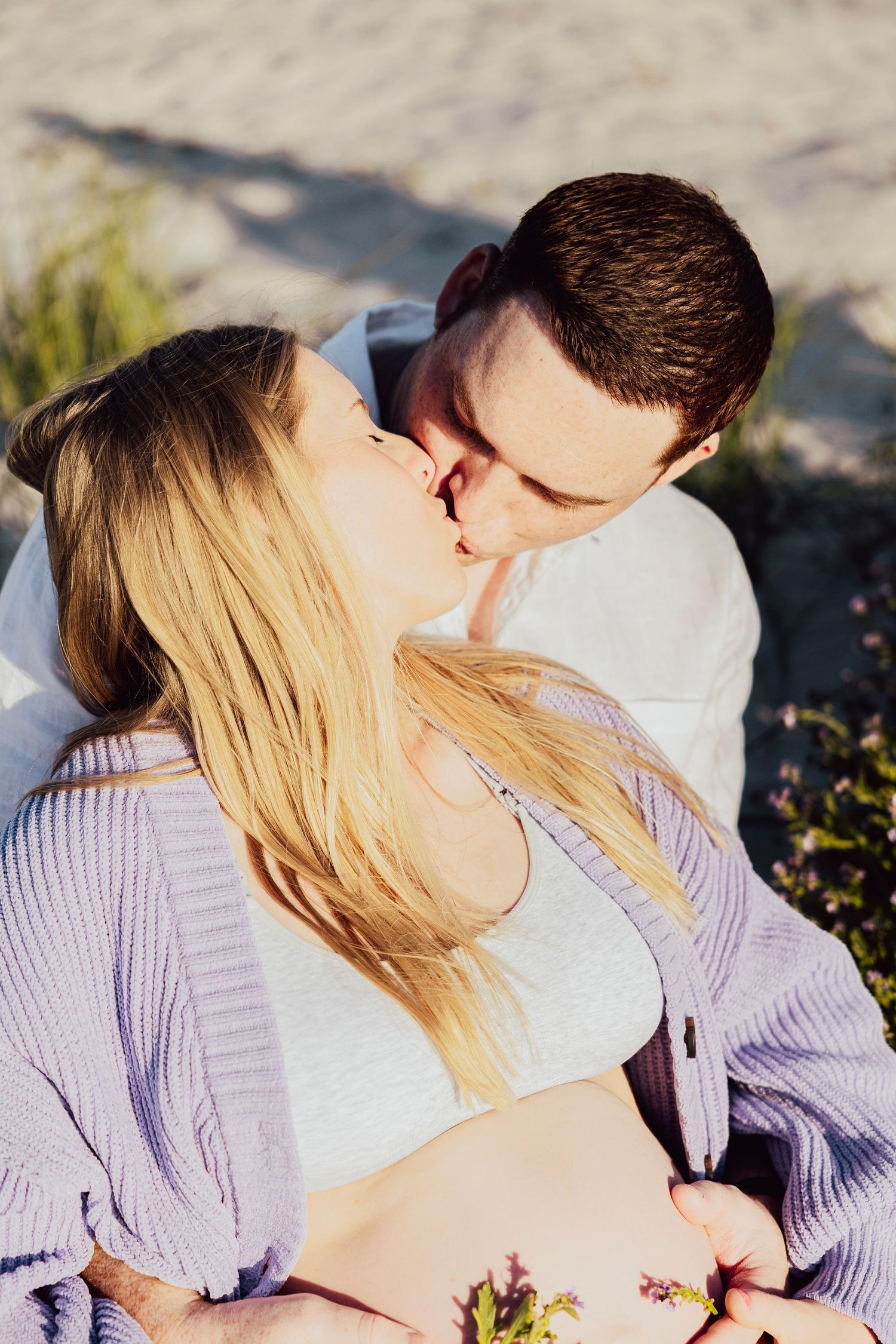 Pregnant couple sharing a kiss while sitting on sandy beach dunes in warm golden sunlight.