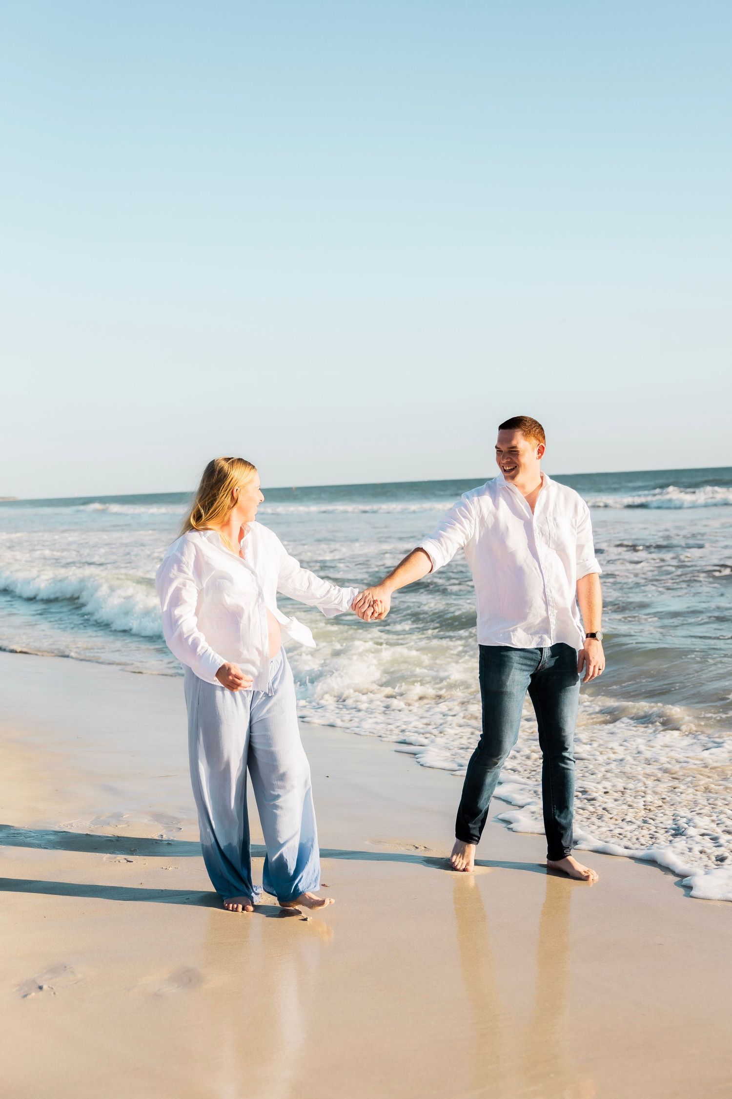 Joyful expecting couple holding hands and walking barefoot along a sunny beach with ocean waves behind them.