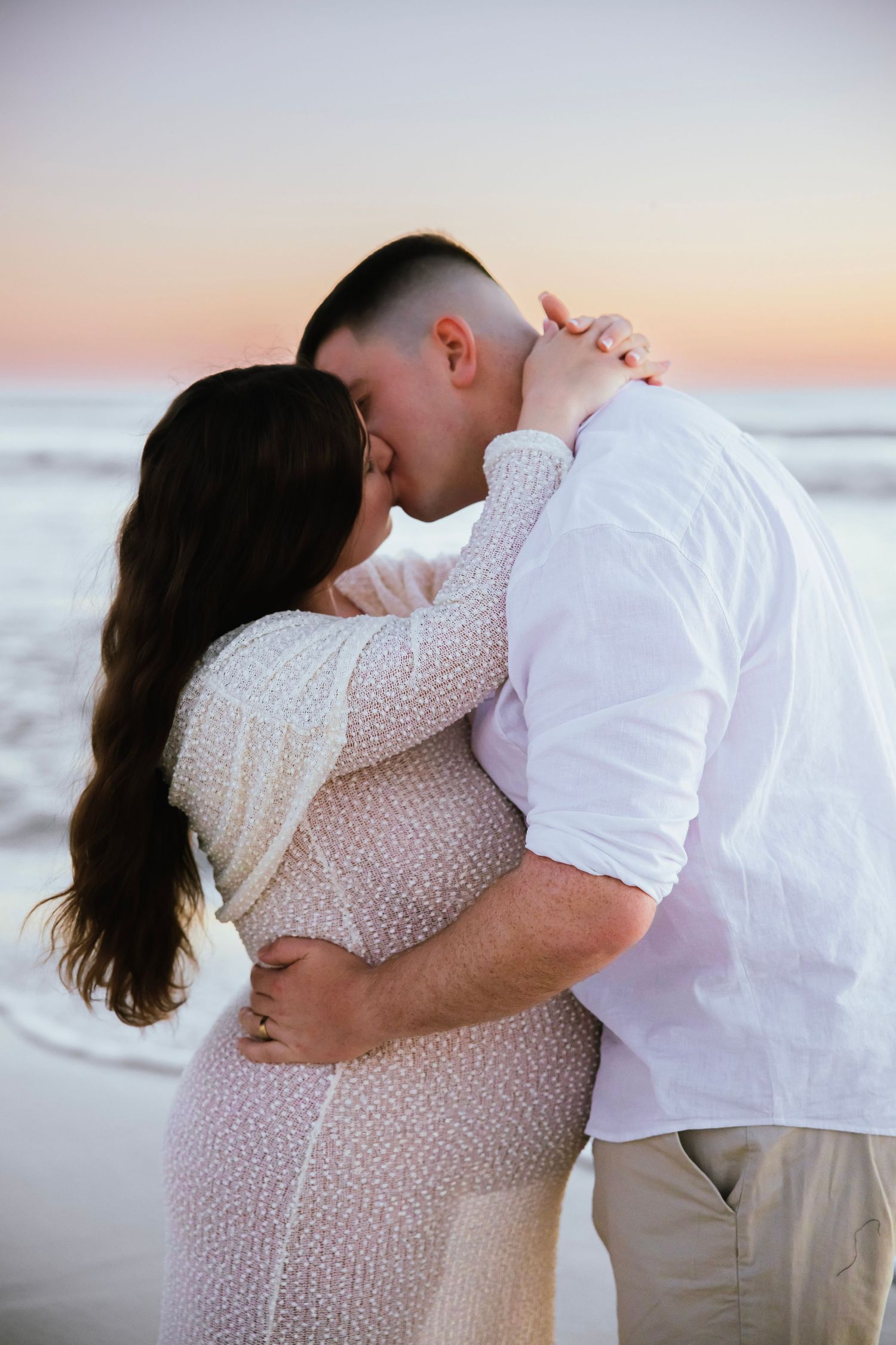 Couple embracing at sunset on beach, man kissing pregnant woman in white lace maternity dress.