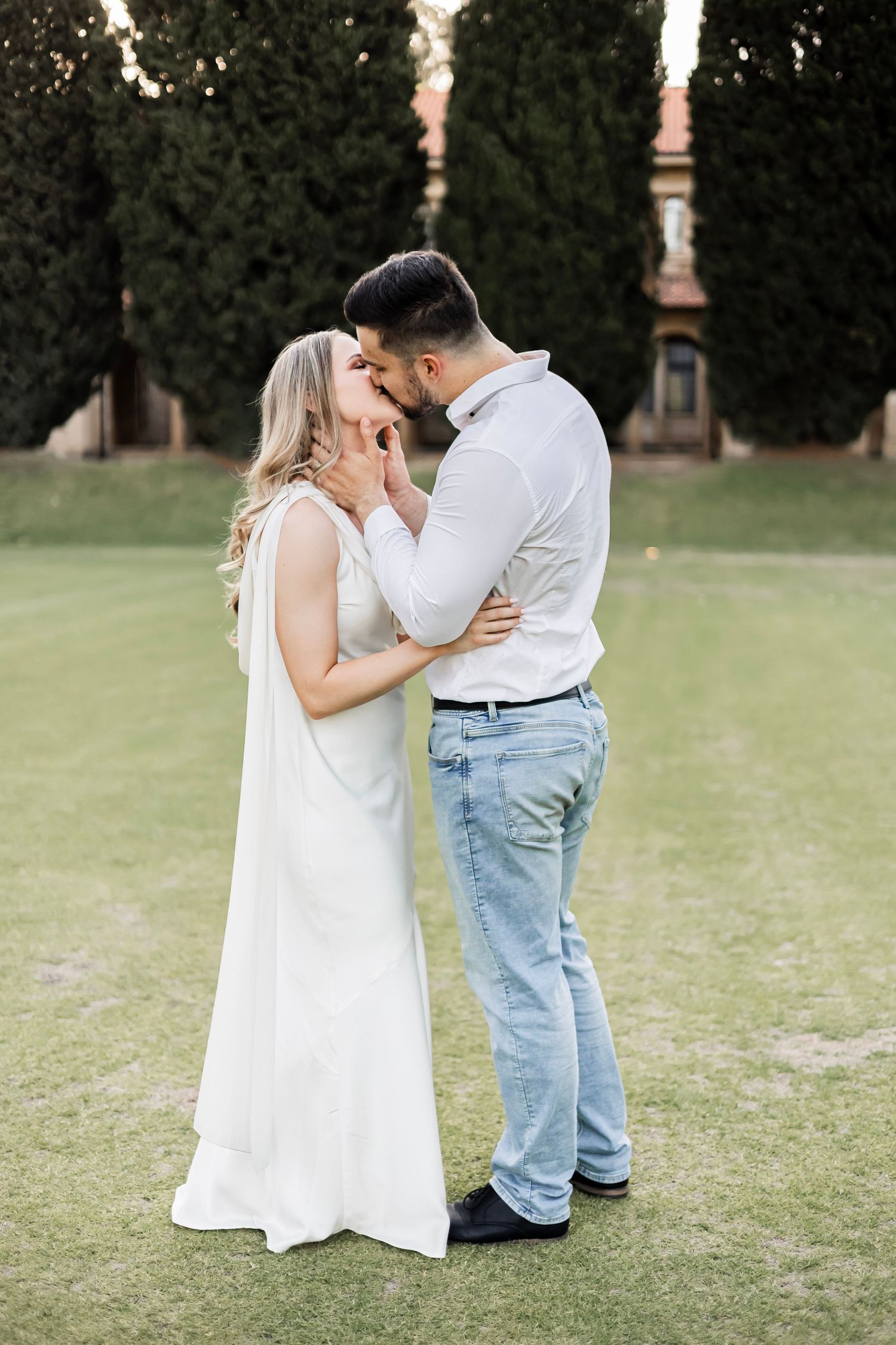 Couple sharing a romantic kiss on green lawn at sunset, bride in white dress embracing groom in white shirt.