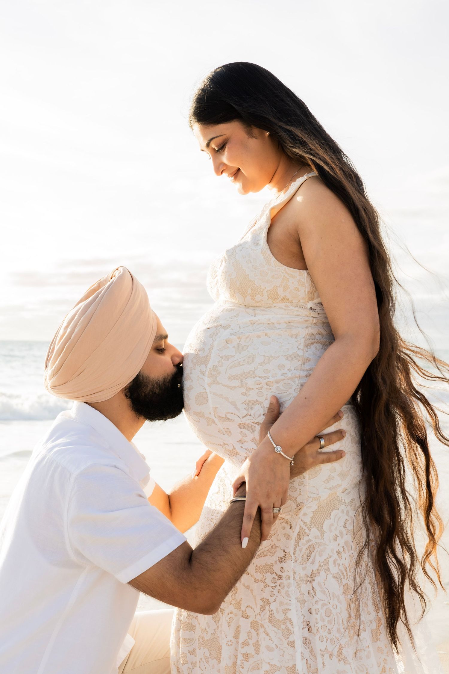 Man in turban kissing pregnant woman's belly on sunny beach, woman smiling down at him tenderly.