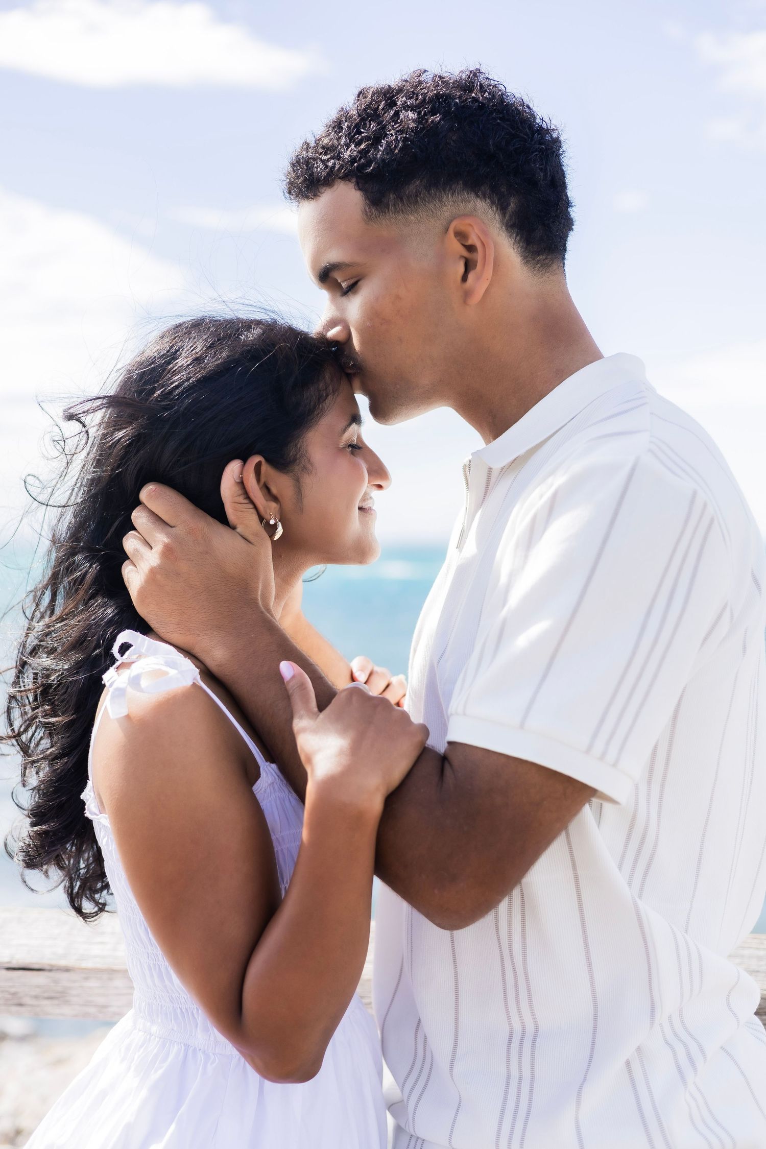 Romantic couple embracing on a sunny beach, man kissing woman's forehead tenderly.