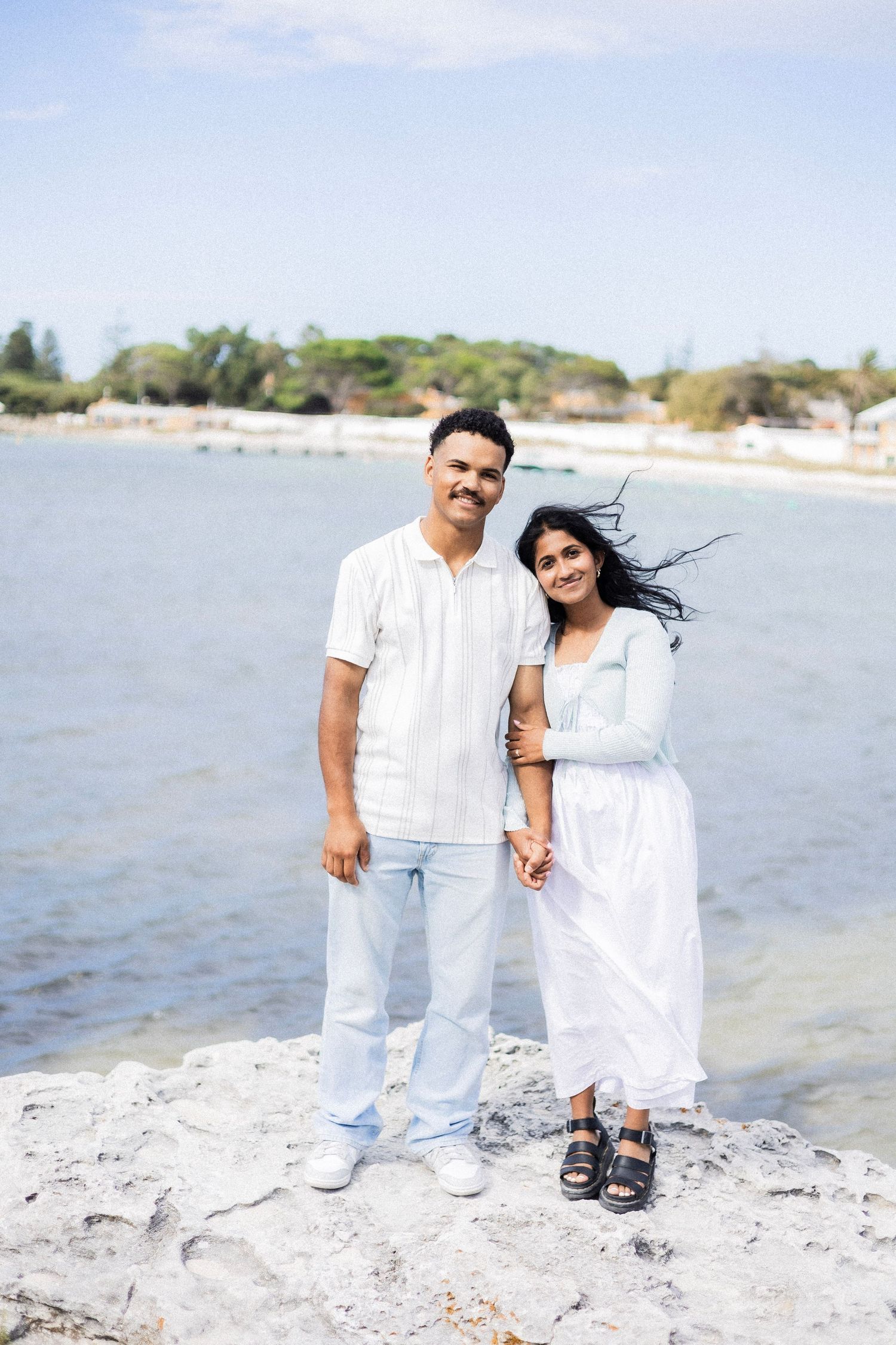A smiling couple stands together on coastal rocks with the ocean and trees in the background.