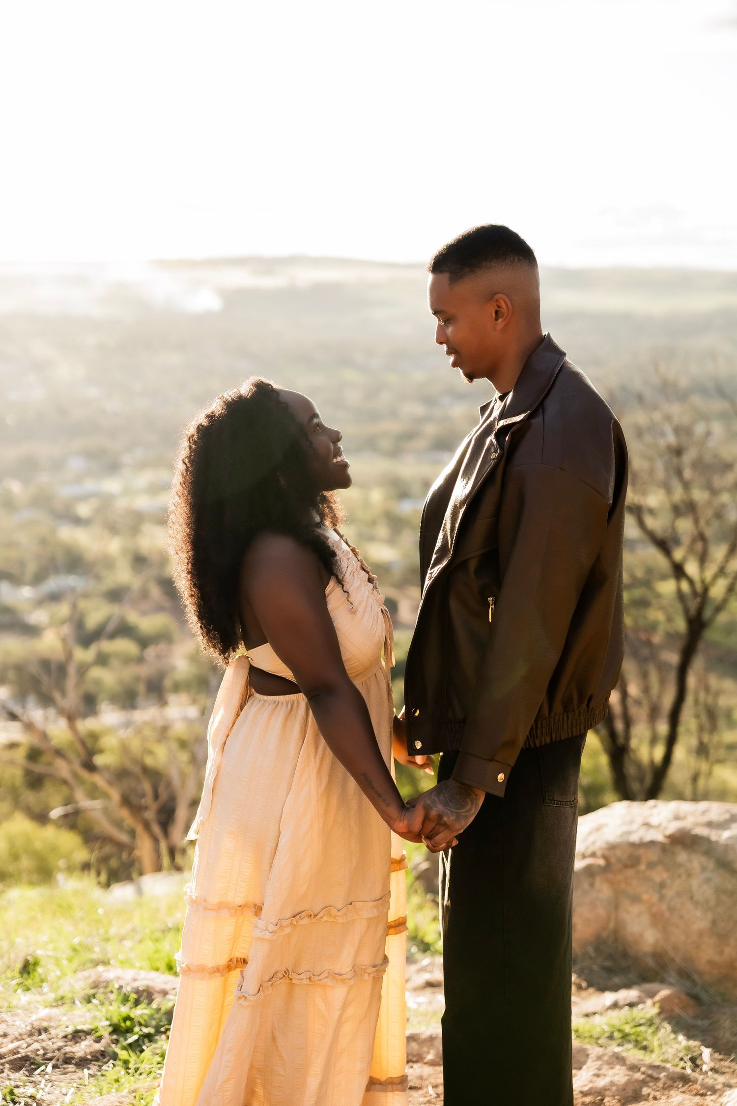 A couple holds hands on a hilltop at golden hour, the woman in a yellow dress gazing up at her partner.
