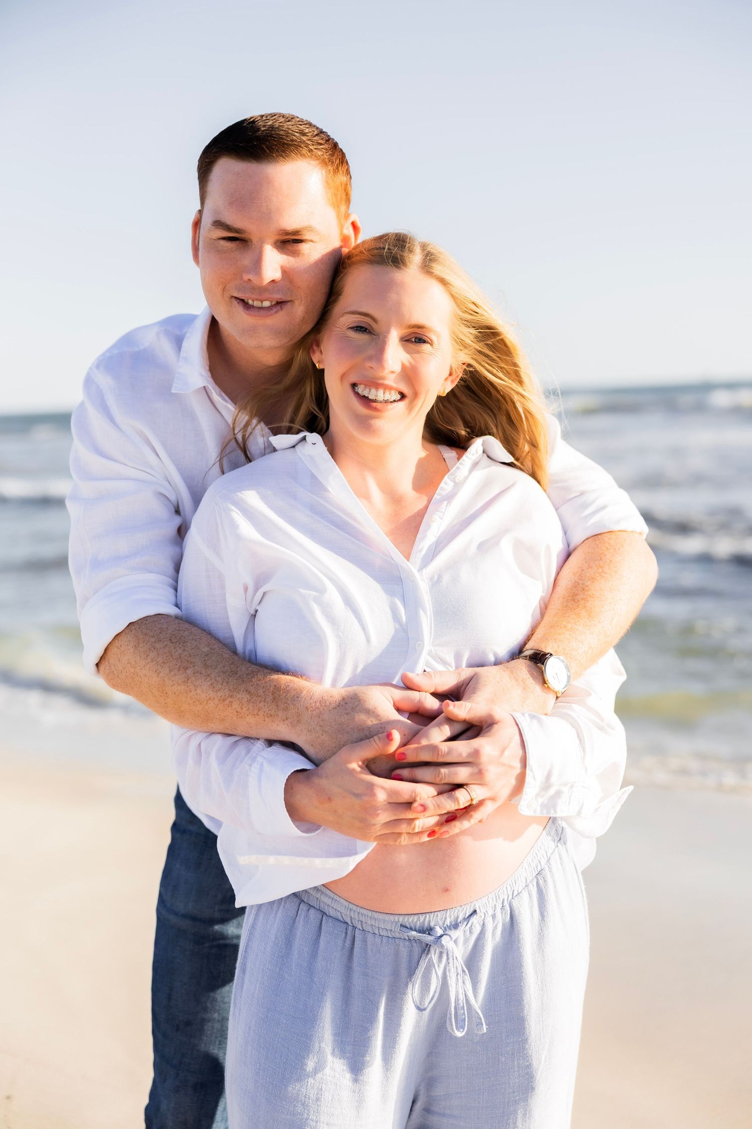Happy couple on beach, man embracing pregnant woman from behind, both smiling with ocean in background.