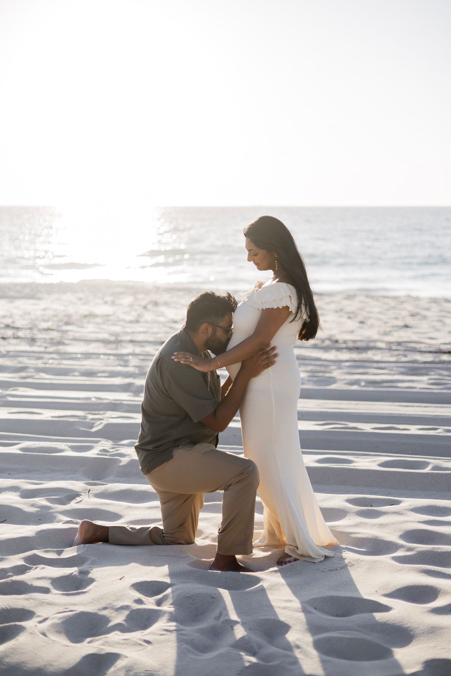 Man kneeling before pregnant woman in white dress on a sun-drenched beach by the ocean.