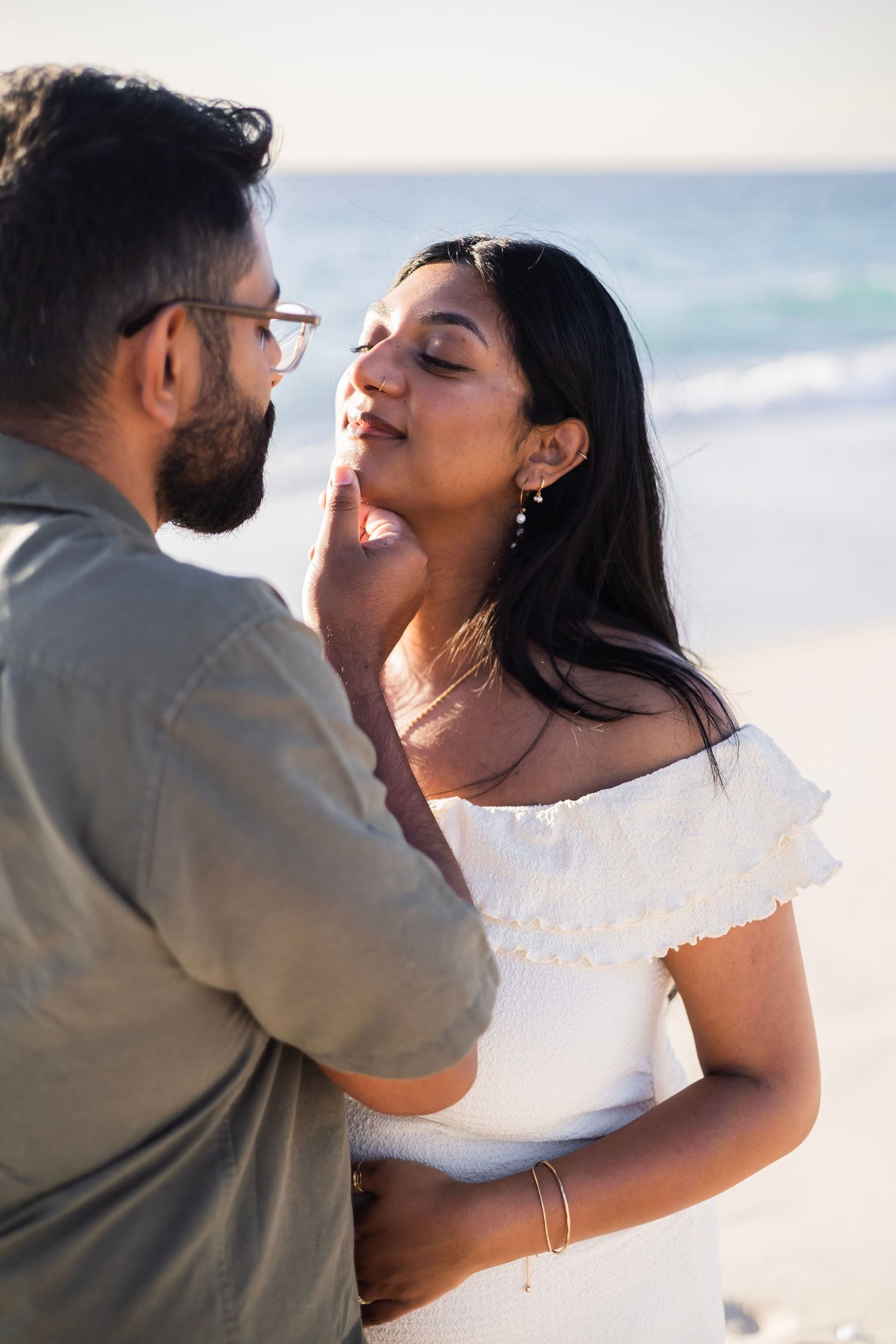 Couple sharing a tender moment on the beach, man gently touching woman's face, woman in white off-shoulder dress.