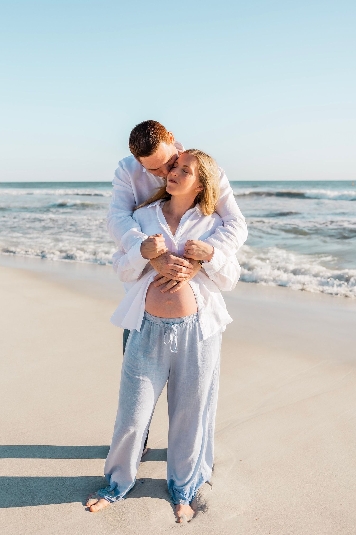 A couple embraces on a sandy beach, the woman visibly pregnant, as gentle ocean waves roll in behind them.