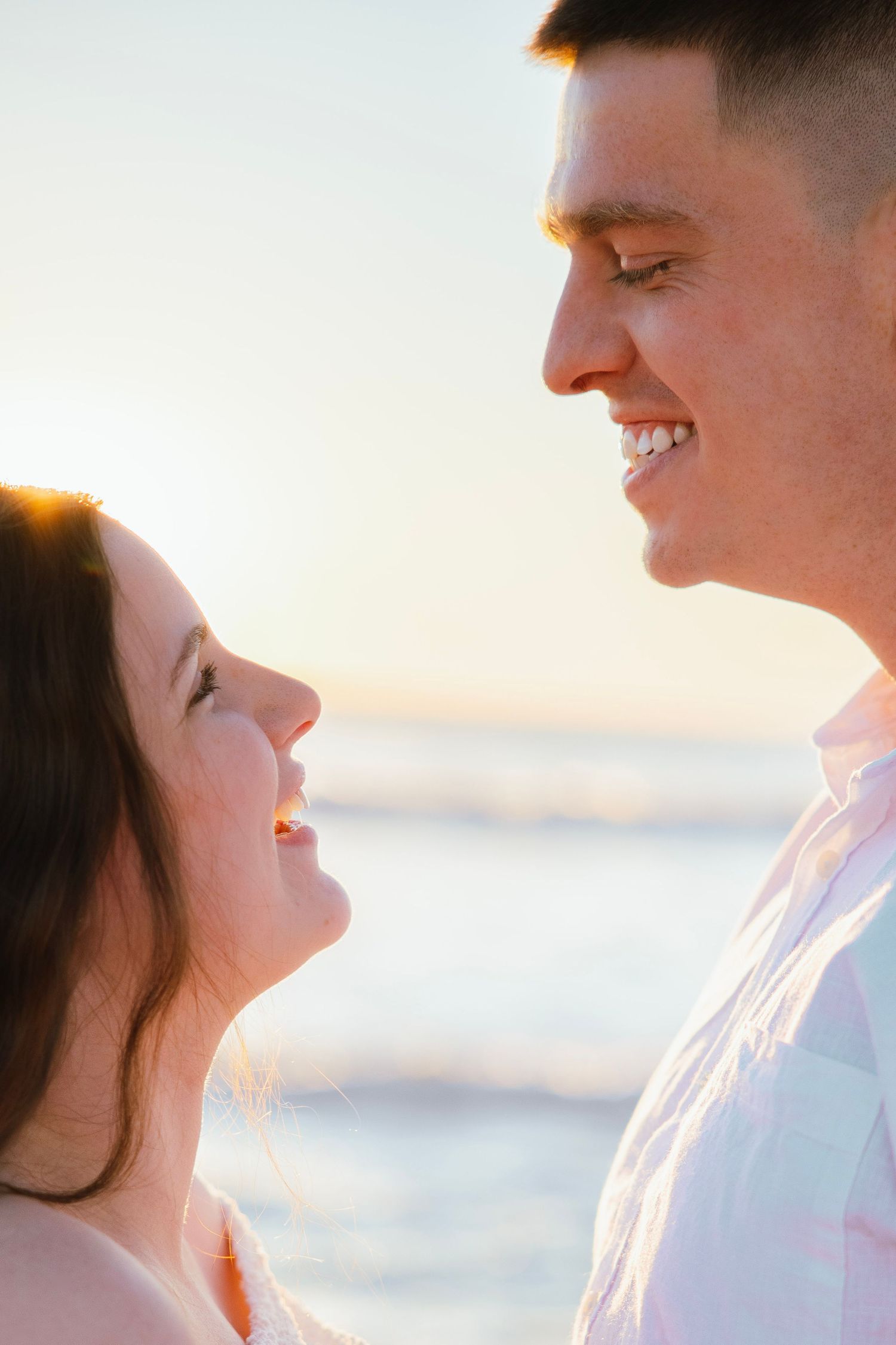 A smiling couple gazes at each other on a sunlit beach at golden hour.