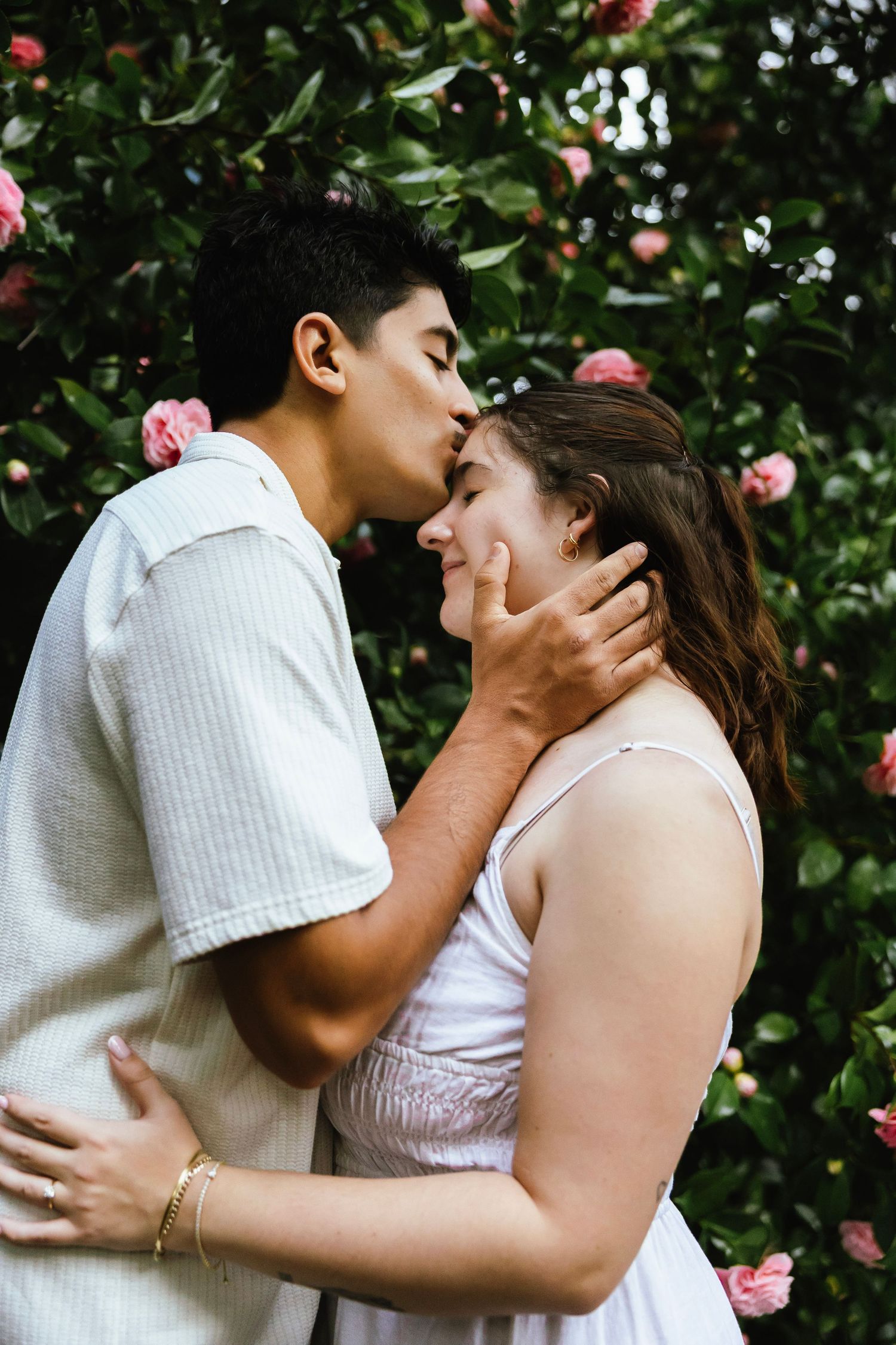 Couple sharing a tender kiss in front of a blooming pink rose bush in a garden.