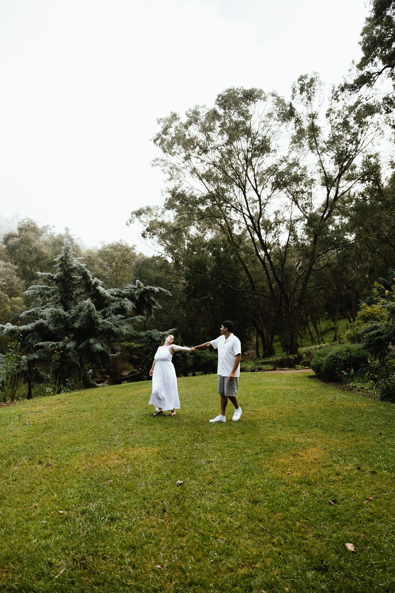 Couple holding hands and dancing on lush green lawn surrounded by tall trees in a park.