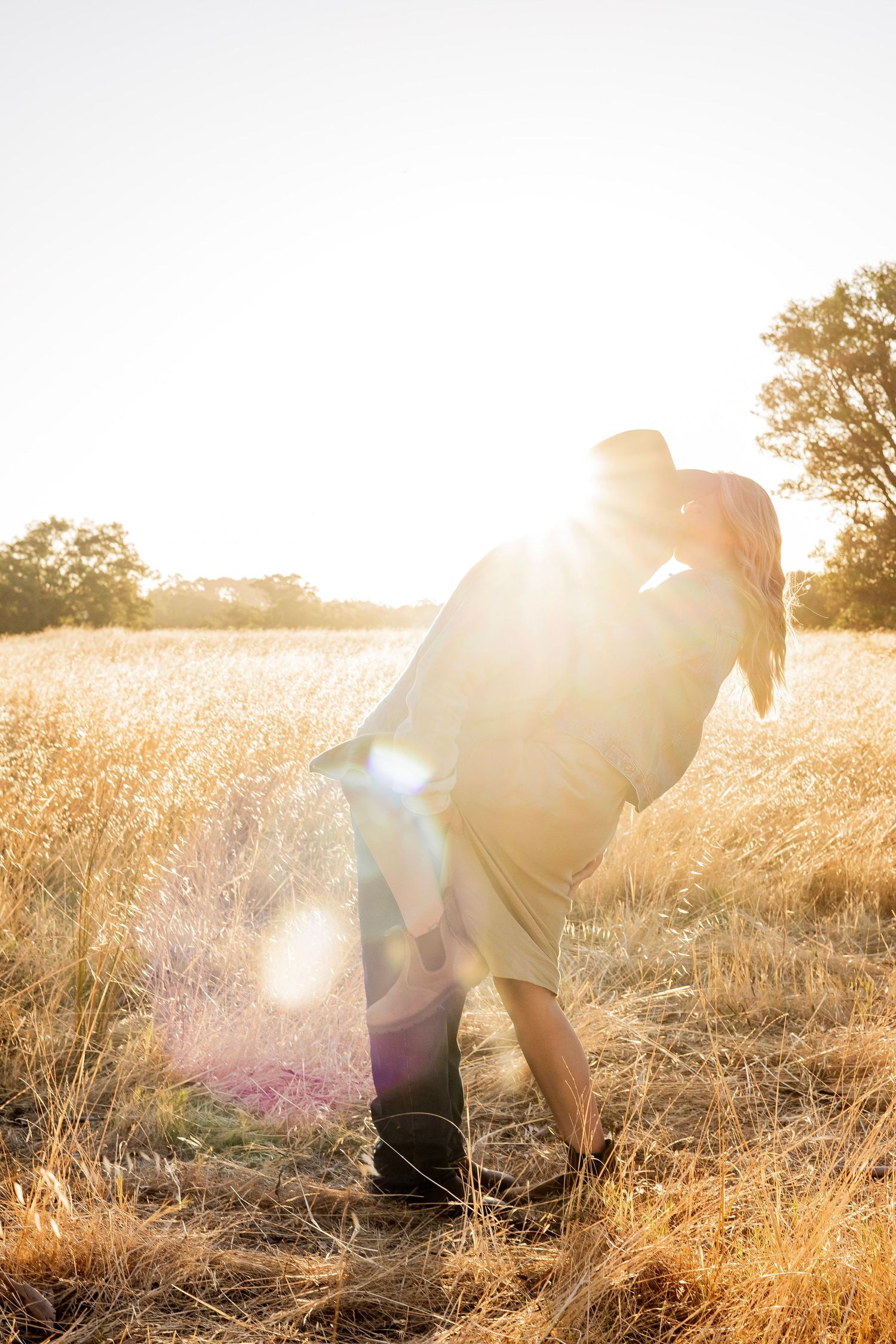 A couple embraces and kisses in a radiant golden field, bathed in warm sunlight with lens flare effects.