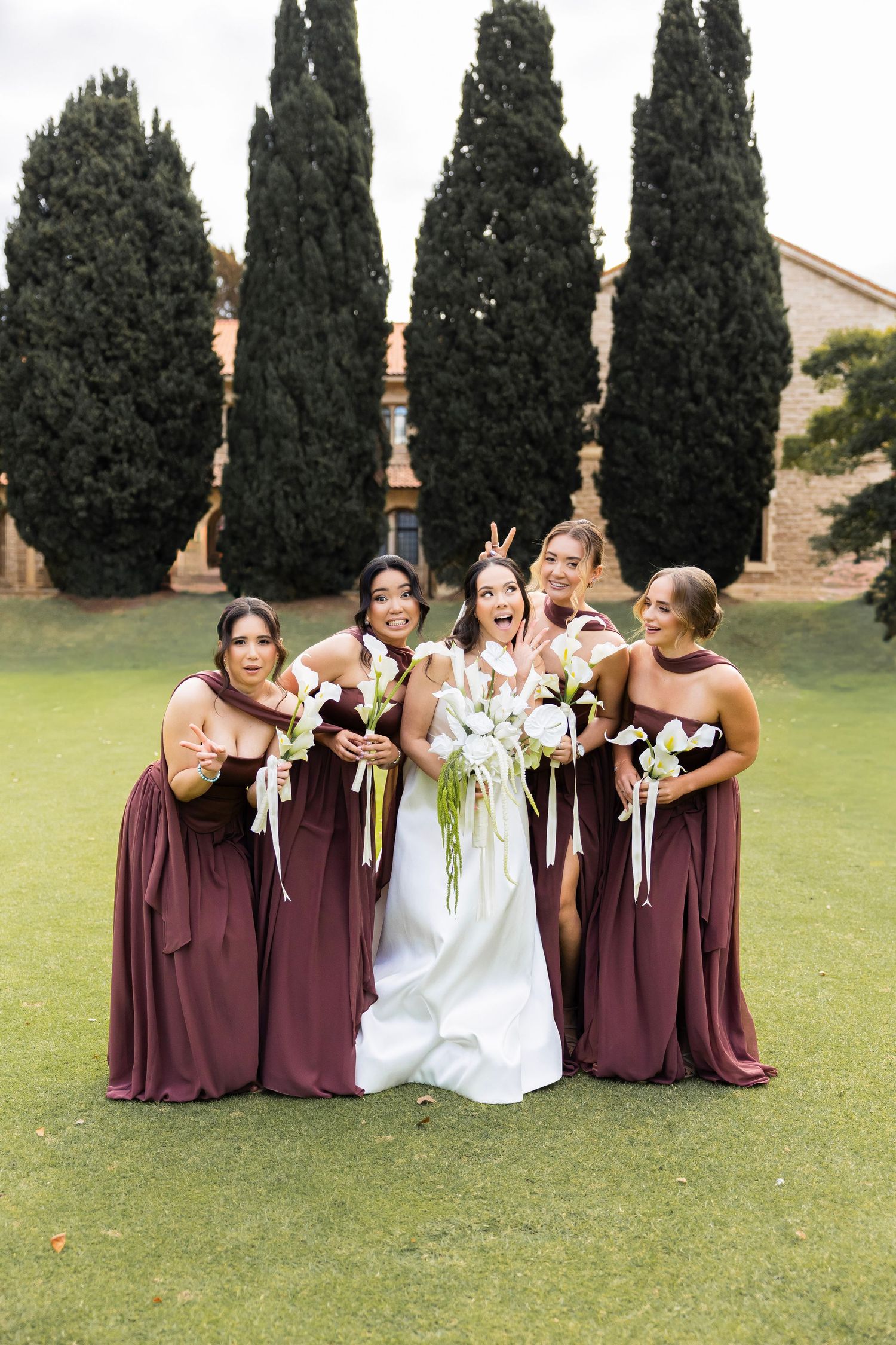 Bride in white gown poses with four bridesmaids in burgundy dresses holding white bouquets on green lawn.