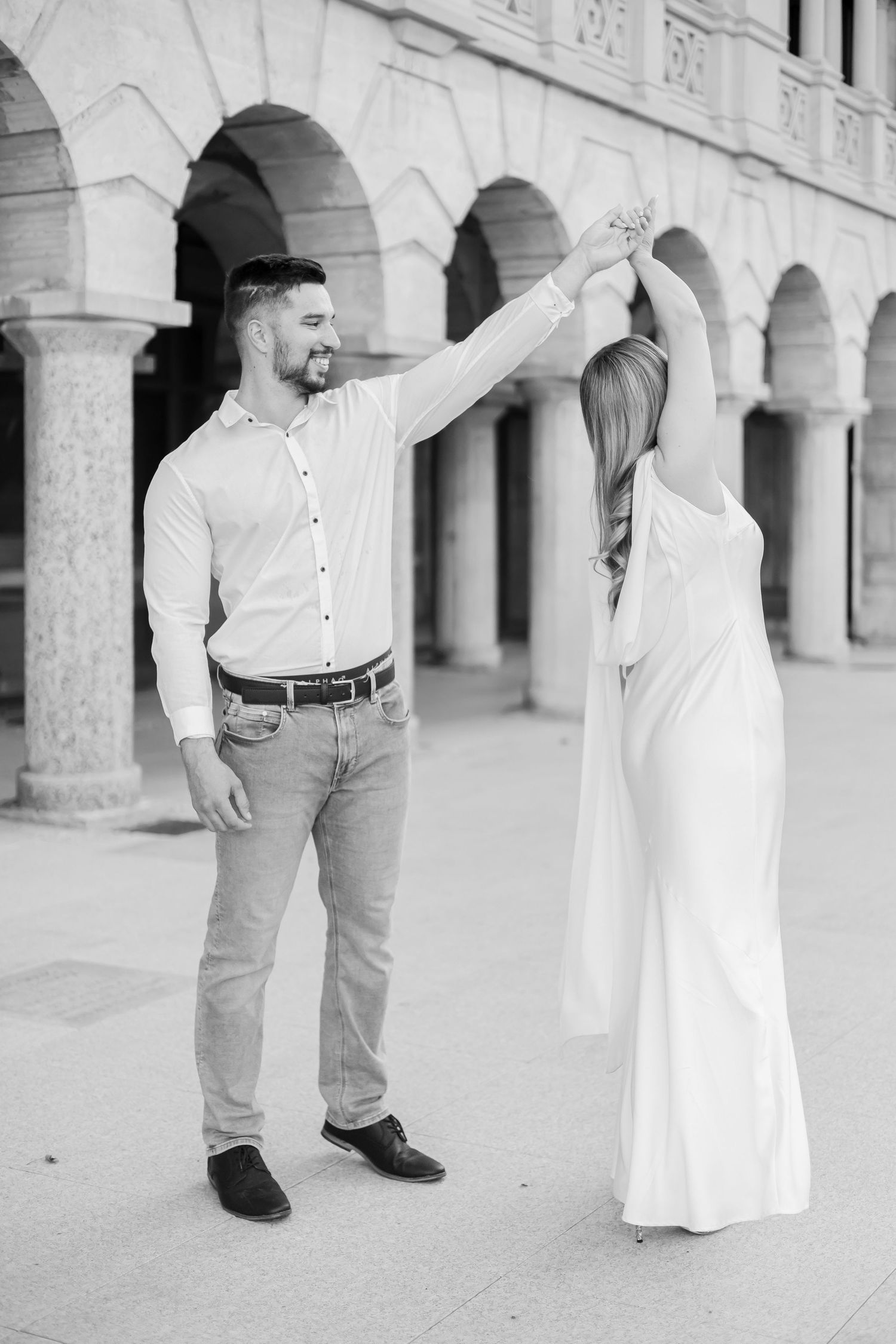 Black and white photo of a couple dancing outdoors, man spinning woman in white dress near arched columns.