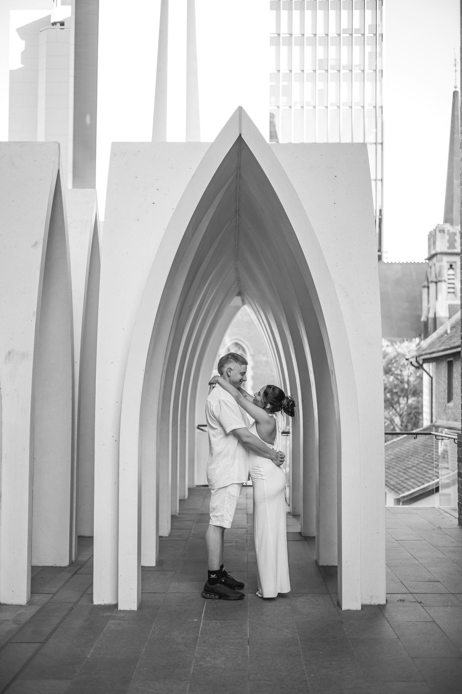 Black and white photo of a couple embracing under gothic arches in an outdoor urban setting.