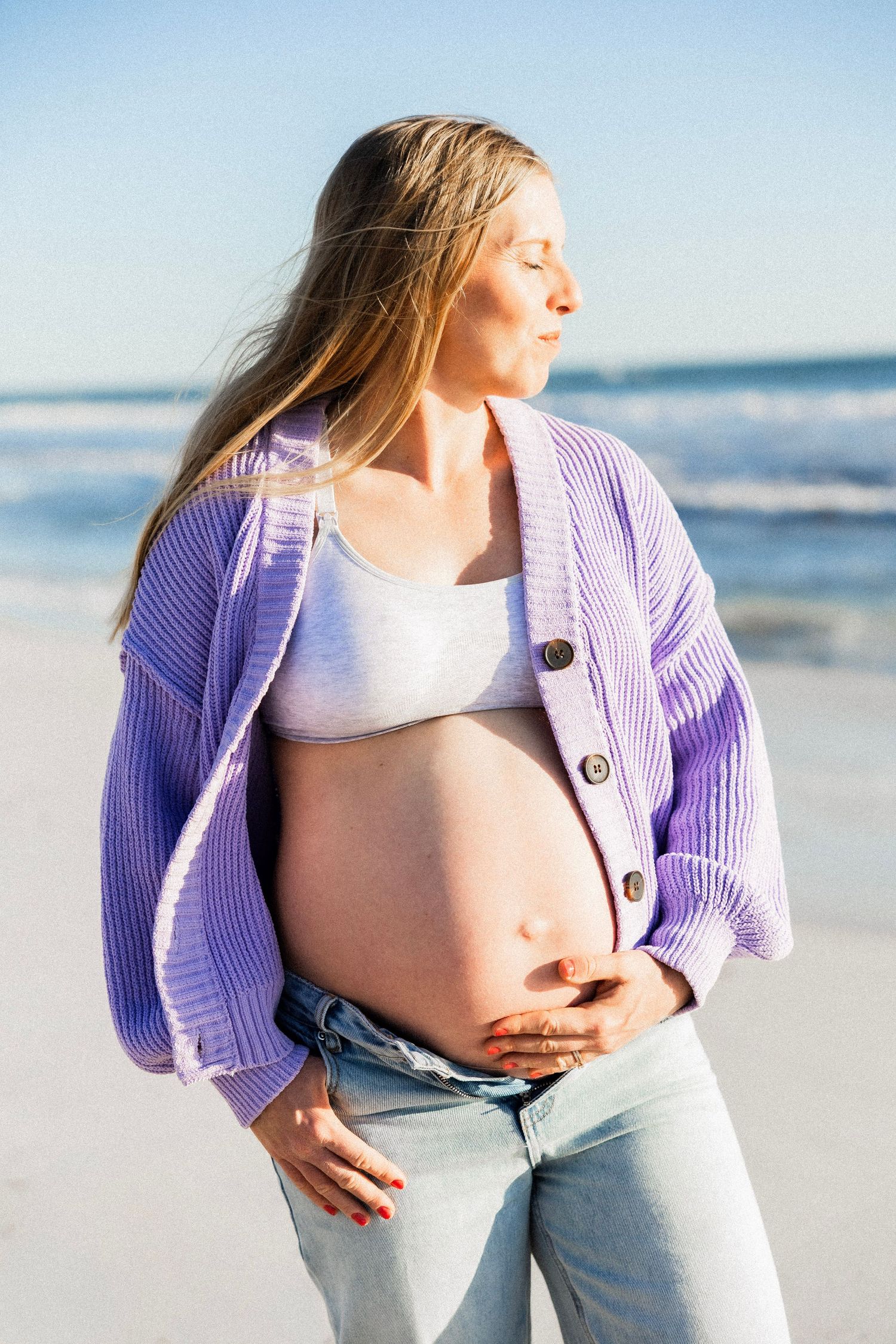 Smiling pregnant woman in purple cardigan and jeans holds baby bump on a sunny beach.