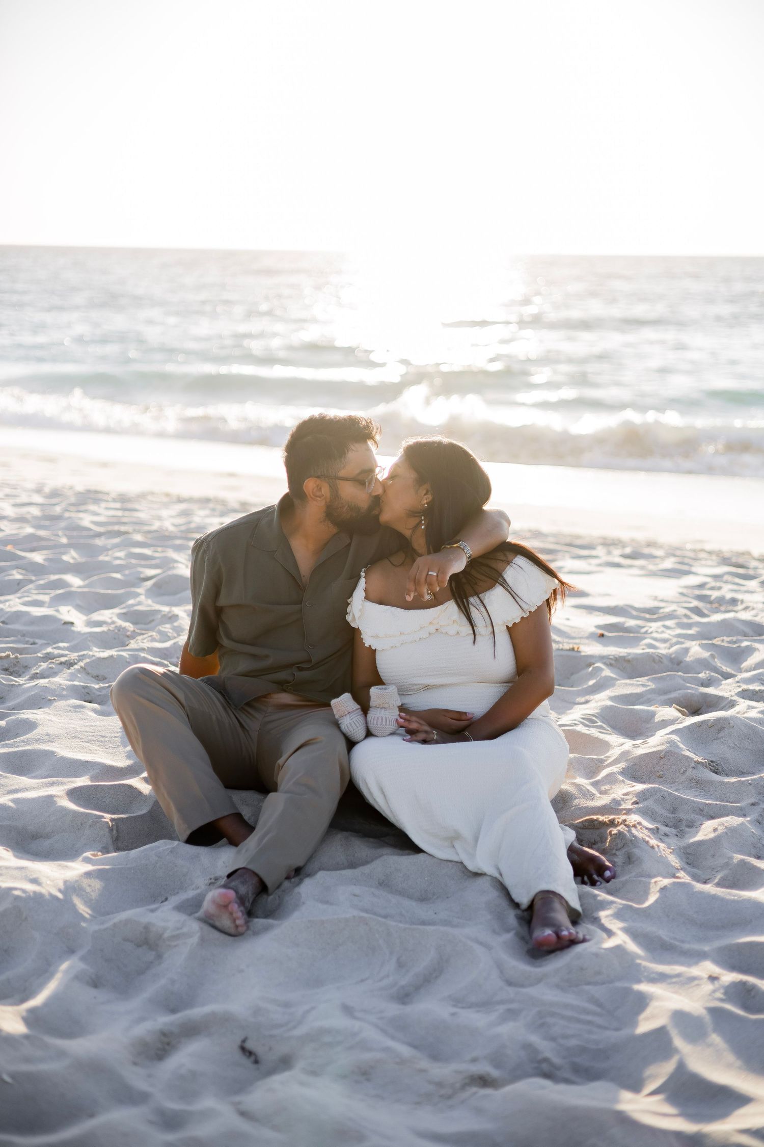 Couple sharing a kiss while sitting on sandy beach at golden hour, woman in white maternity dress.