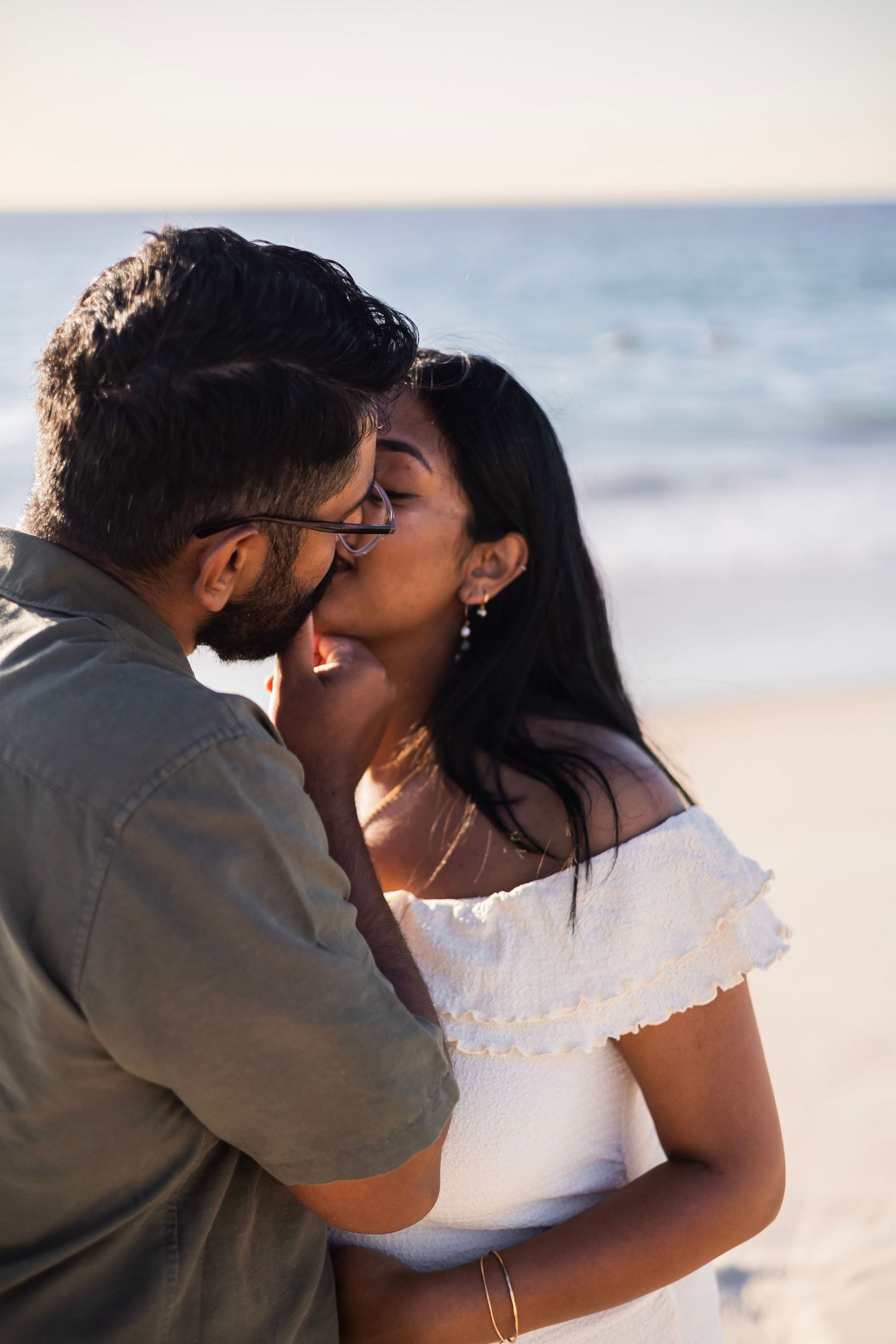 A romantic couple sharing a tender kiss on a sunny beach, woman in white off-shoulder dress, ocean in background.