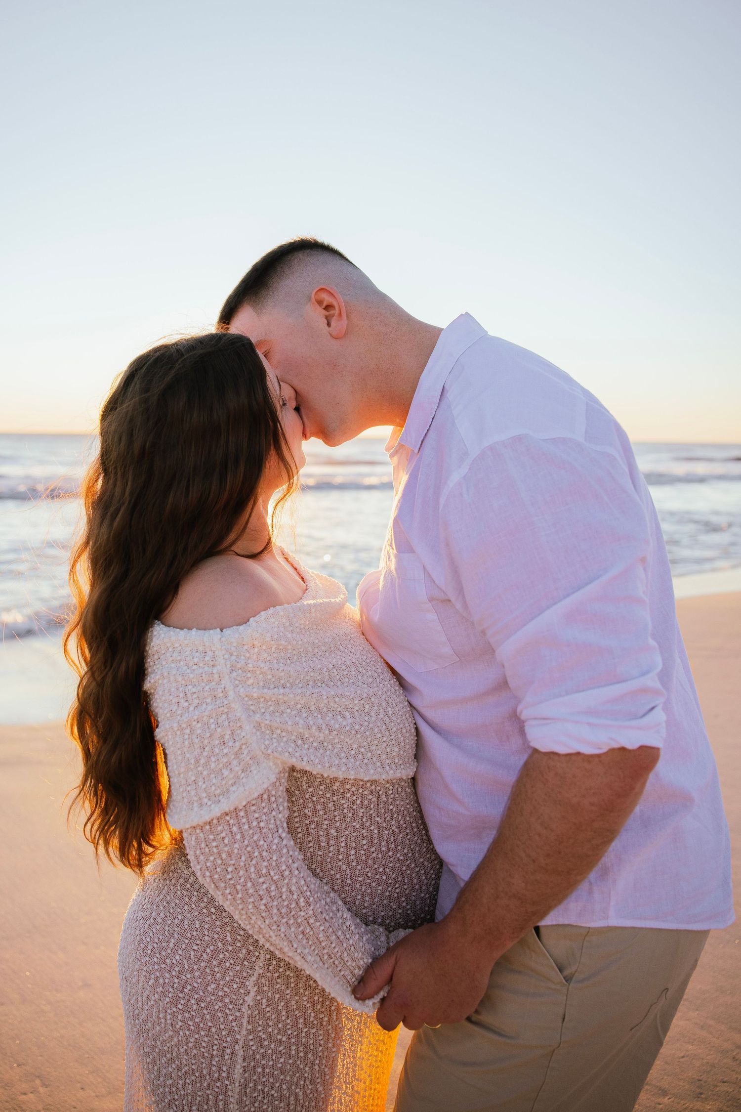 Man kissing pregnant woman's head on beach at sunset, both dressed in white, holding her baby bump.