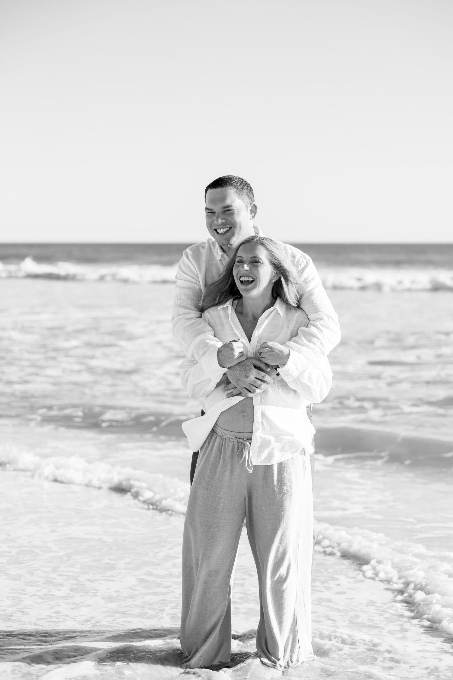 Black and white photo of a happy couple embracing and laughing together on a sunny beach with ocean waves.