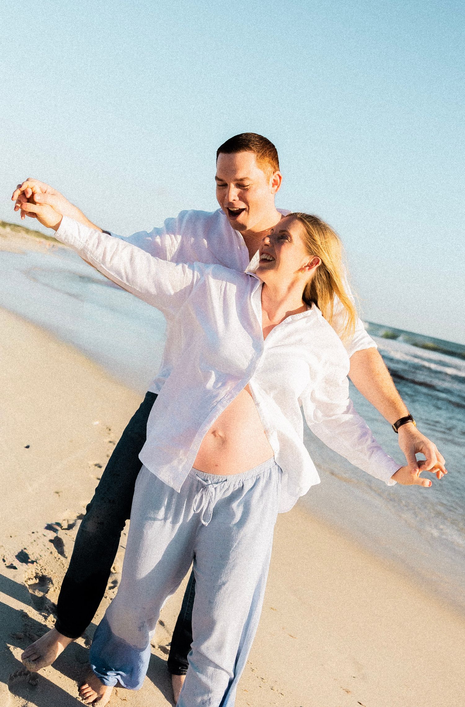 Joyful pregnant couple dancing on a sunny beach, wearing white shirts, with ocean waves in the background.