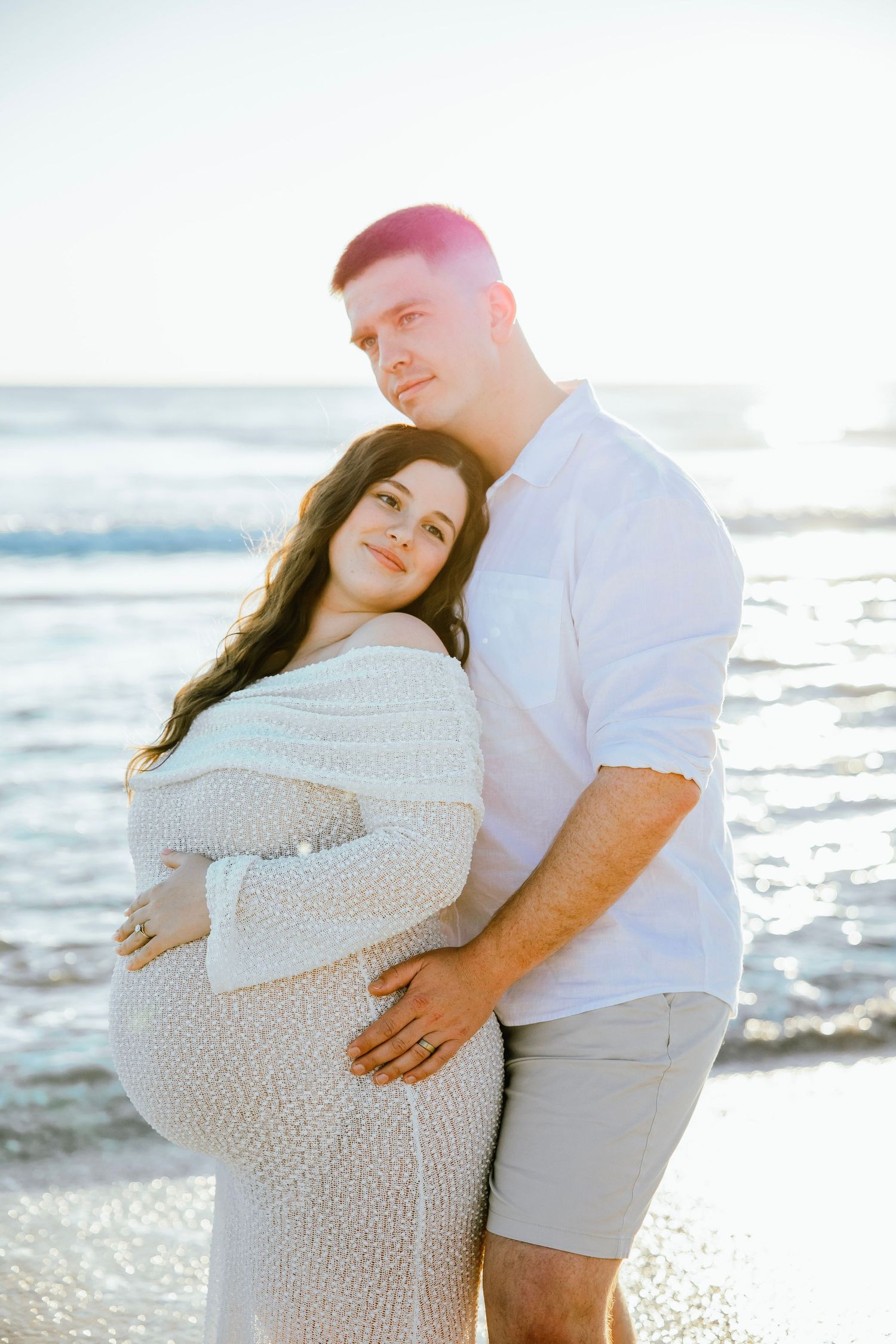 Pregnant couple embracing on beach at sunset, woman in white knit dress cradling baby bump, ocean waves behind them.