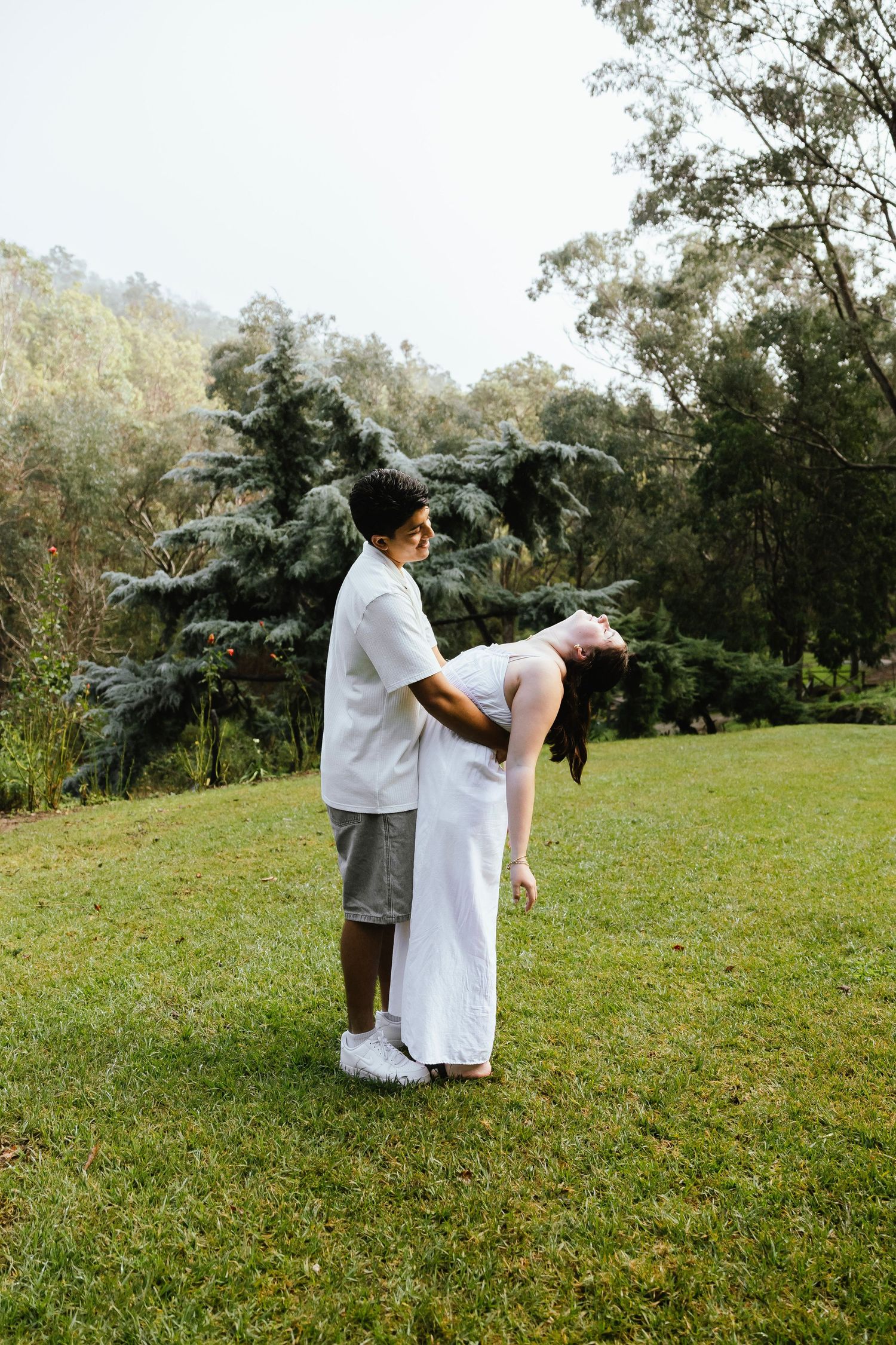 Couple dancing outdoors on green grass, woman in white dress leaning back, surrounded by lush trees in a garden setting.