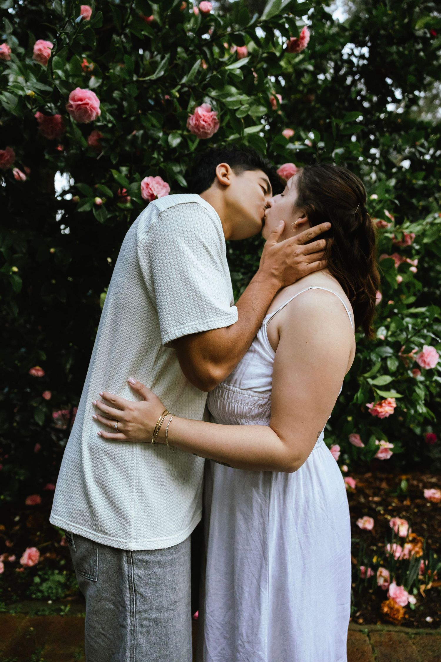 Couple sharing a romantic kiss in front of a blooming pink rose bush, dressed in casual white outfits.