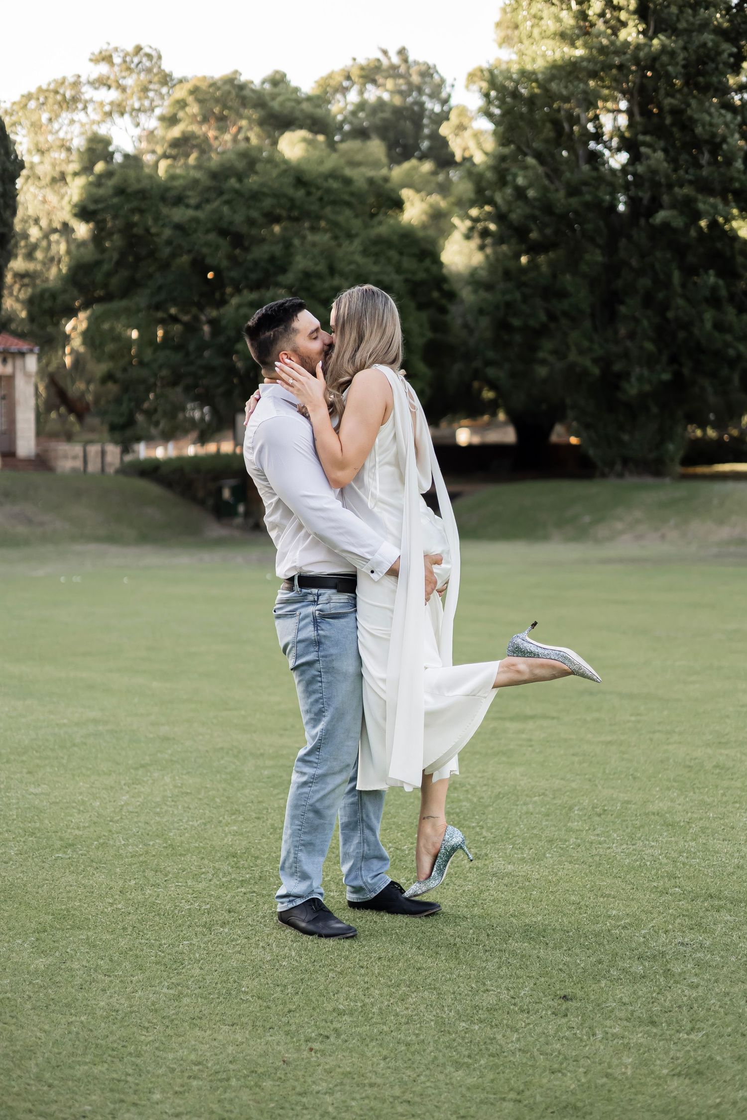 Couple sharing a romantic kiss on a lush green field, woman in white dress lifting her leg, trees in background.