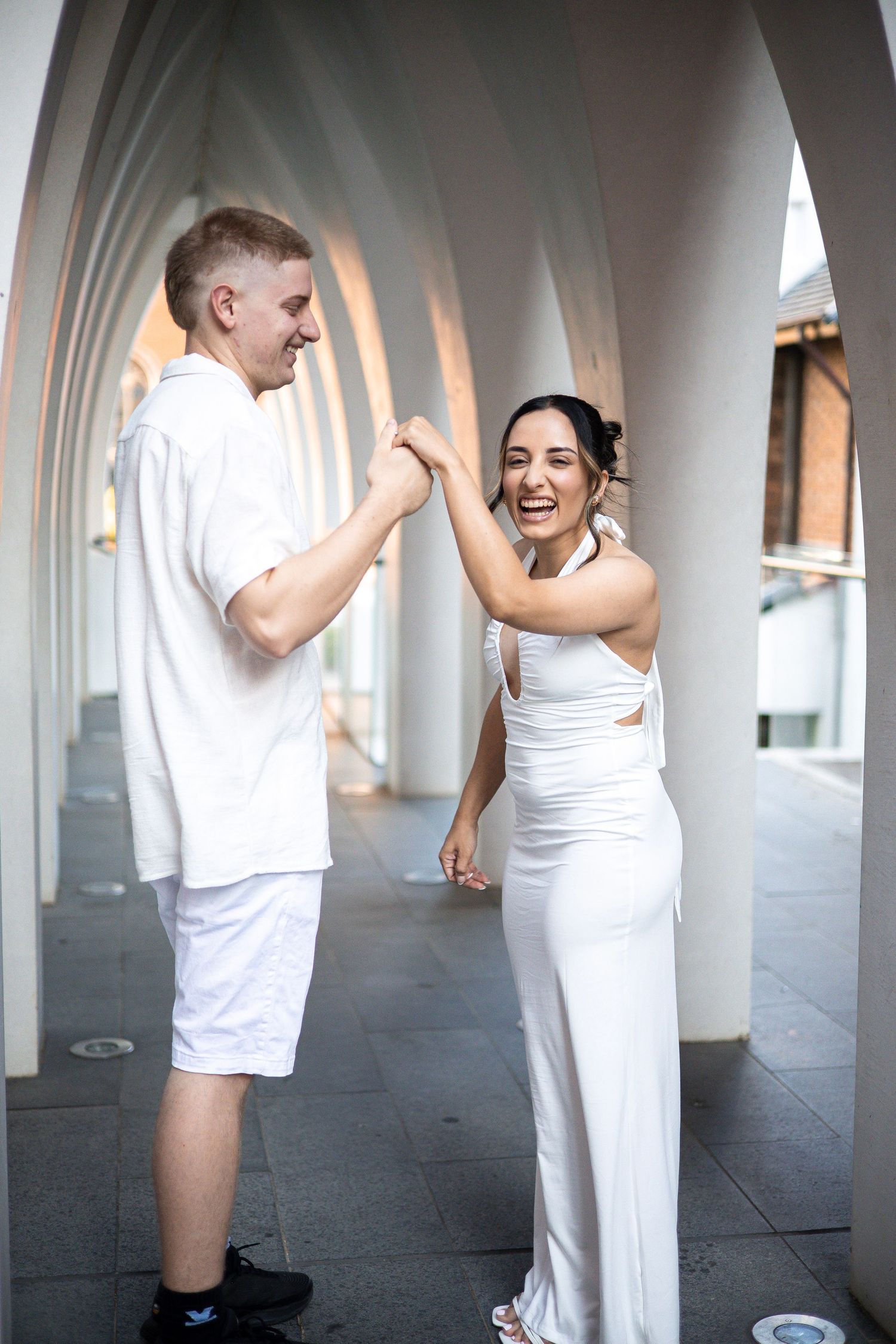Happy couple in white outfits dancing under elegant arched colonnade during engagement photo session.