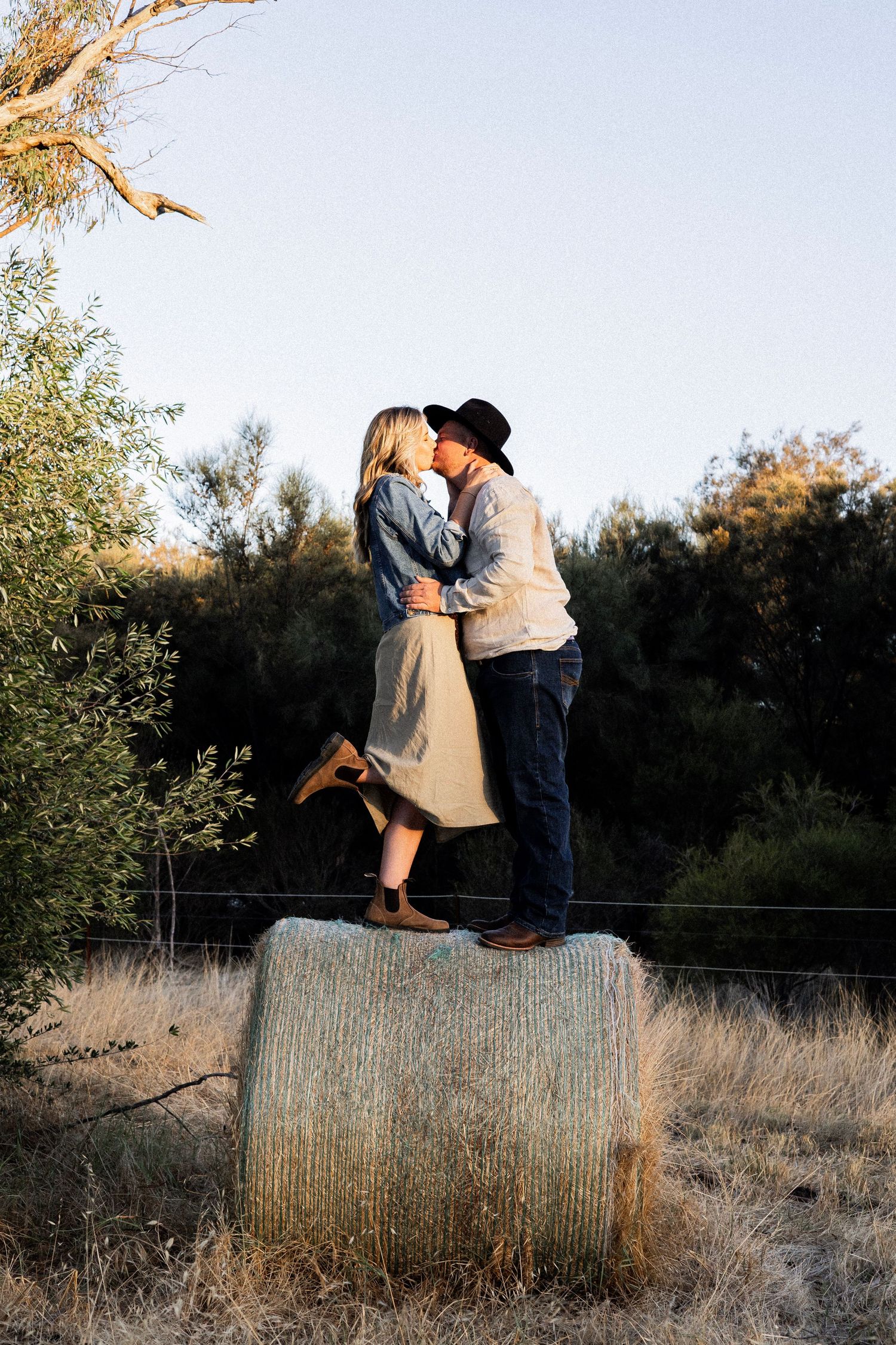 Couple kissing on top of a hay bale at sunset, surrounded by trees in a rural countryside setting.