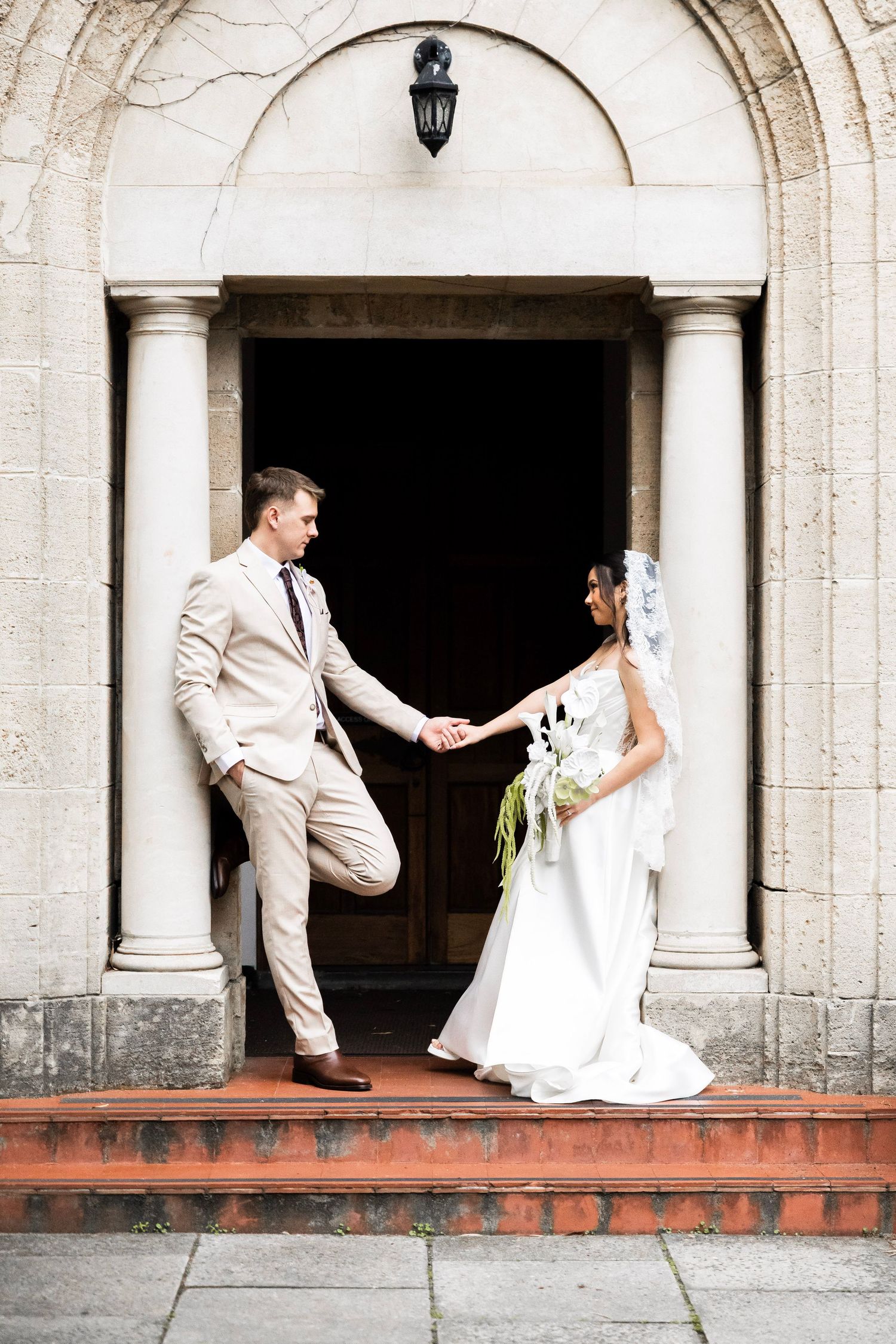 Bride and groom hold hands on church steps beneath stone archway, bride carrying white floral bouquet.