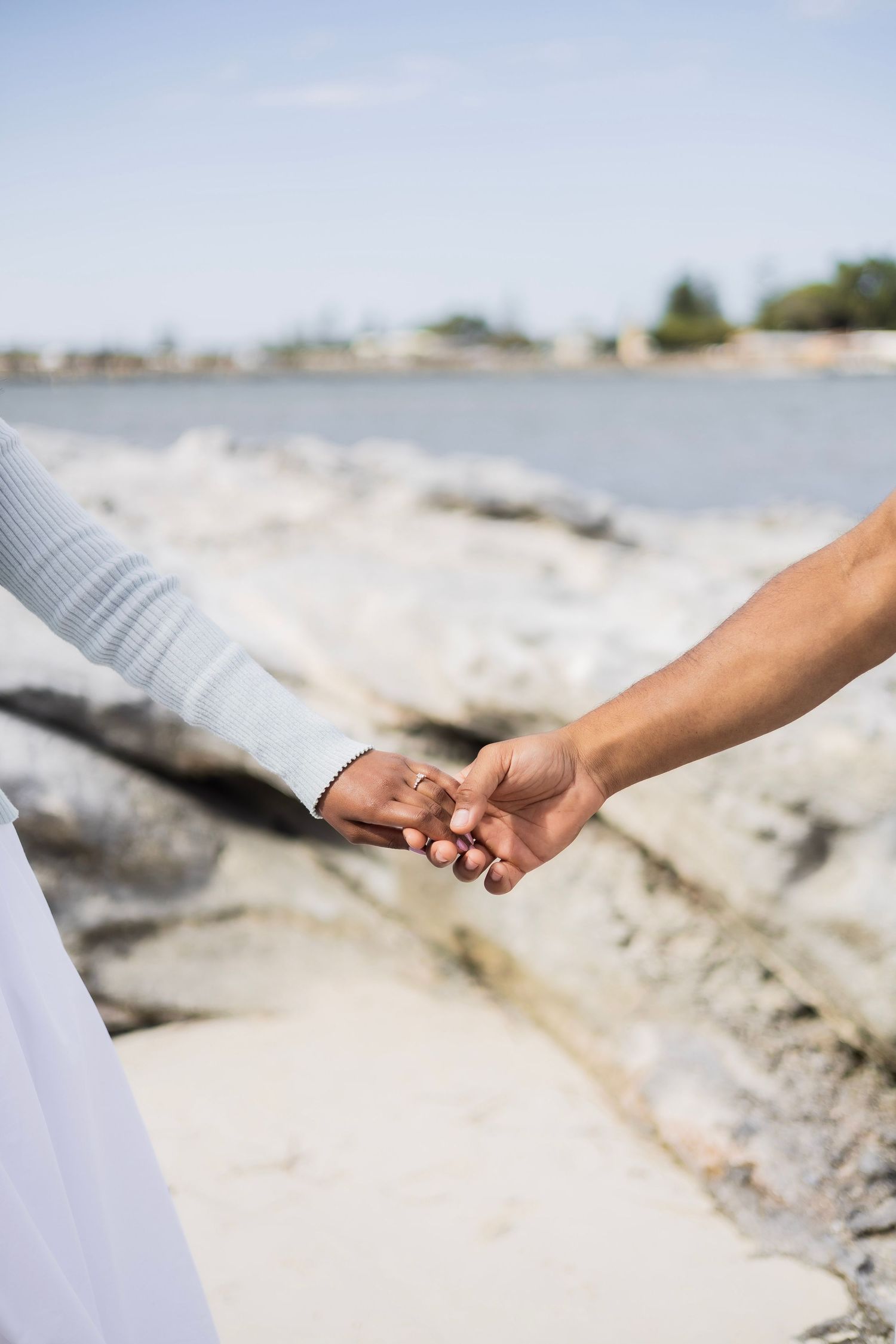 Close-up of an interracial couple holding hands on a rocky beach with blue sky and water in the background.