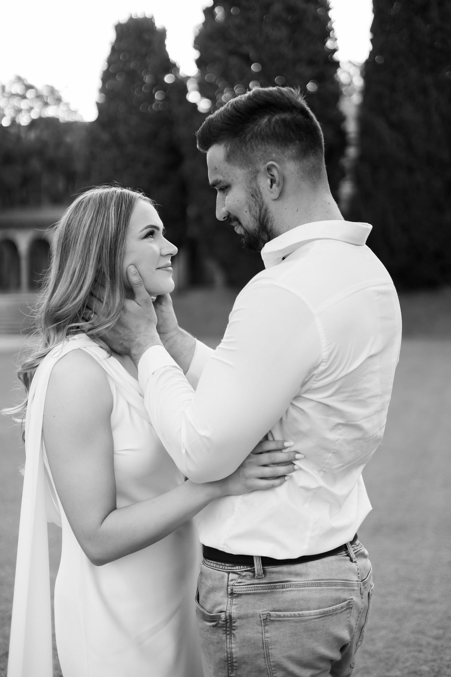 Black and white photo of romantic couple gazing at each other outdoors near cypress trees.