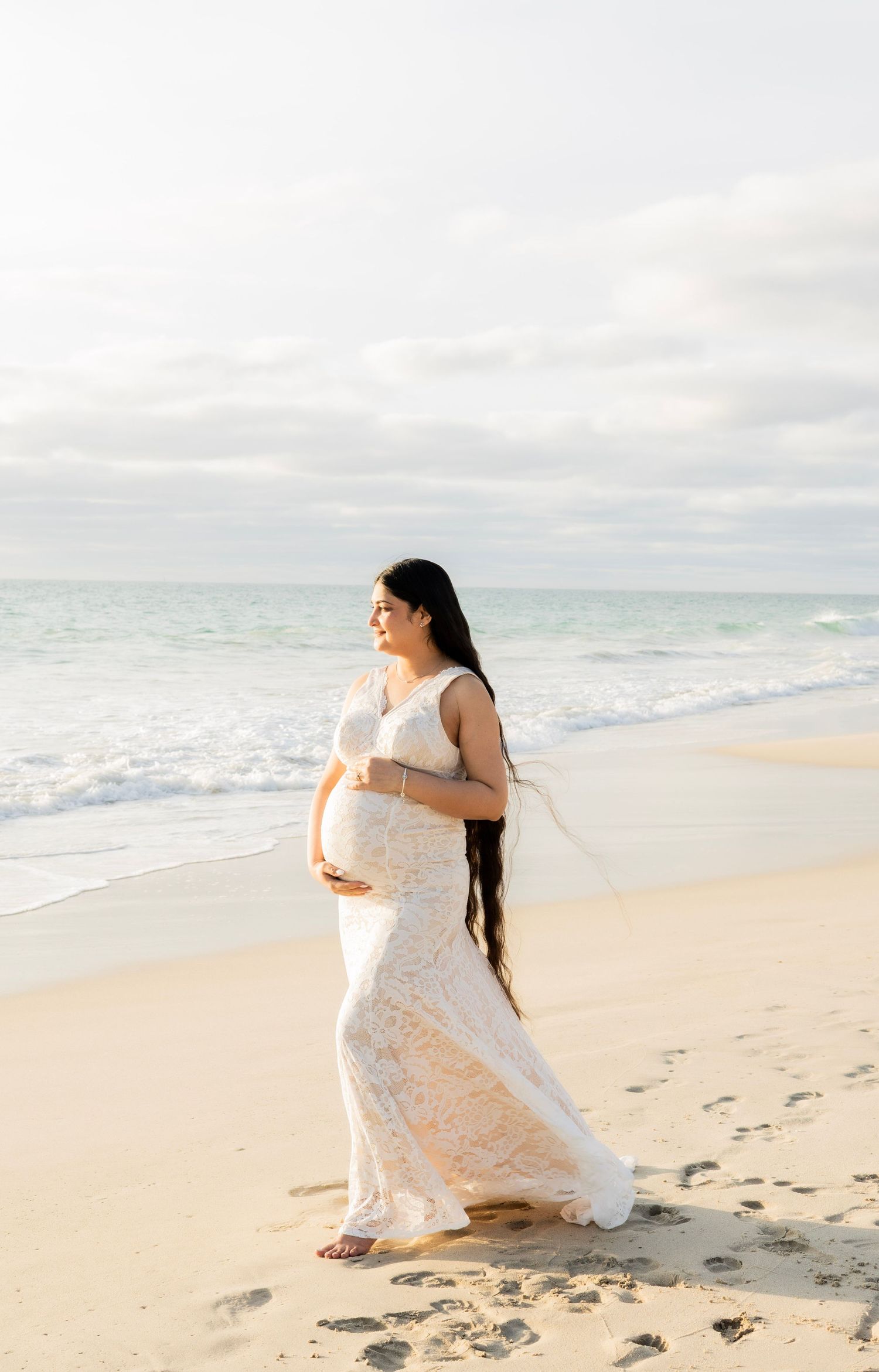 Glowing pregnant woman in white lace dress standing on sandy beach, holding belly near ocean waves.
