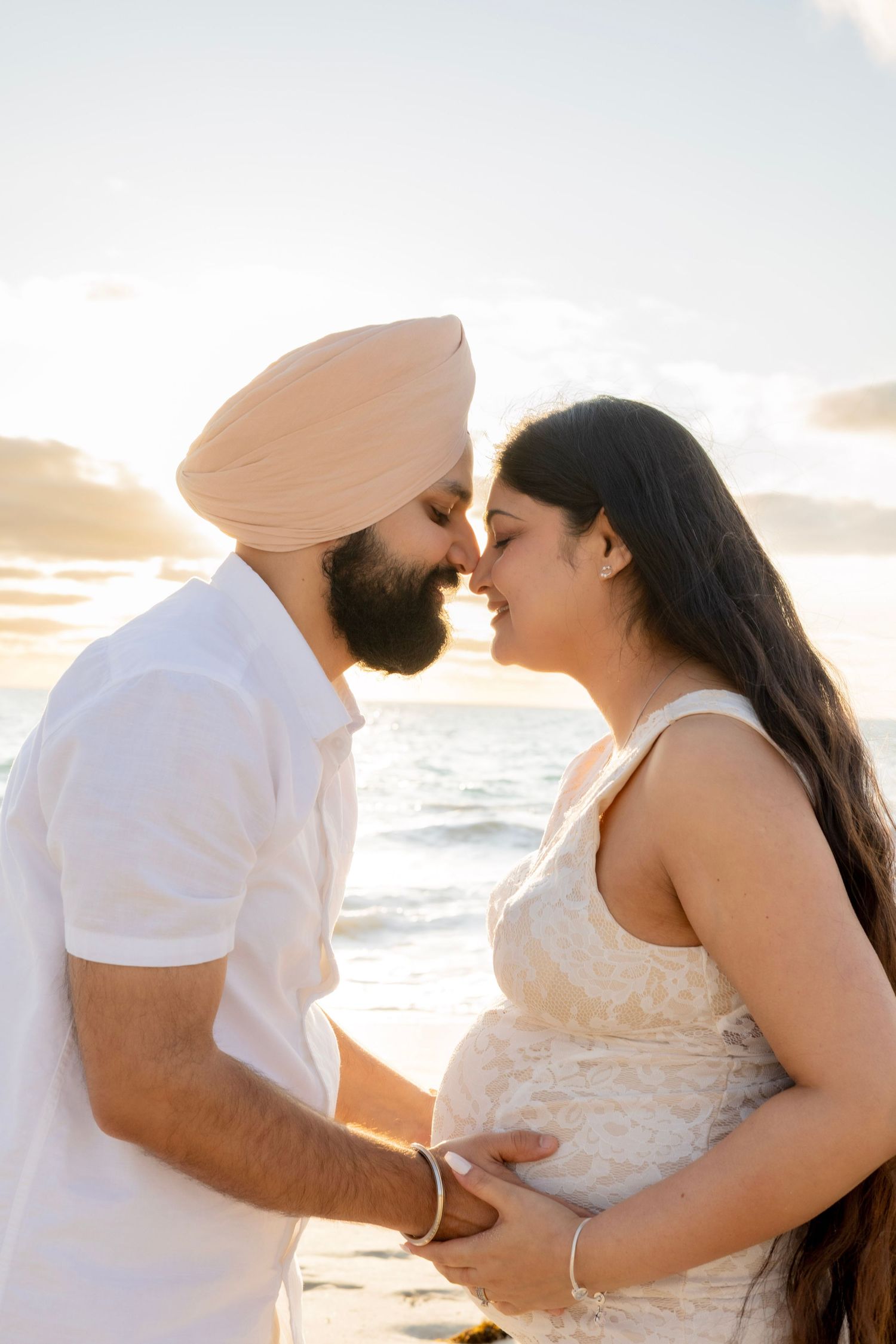 Pregnant couple sharing an intimate moment at sunset on the beach, man wearing a pink turban.