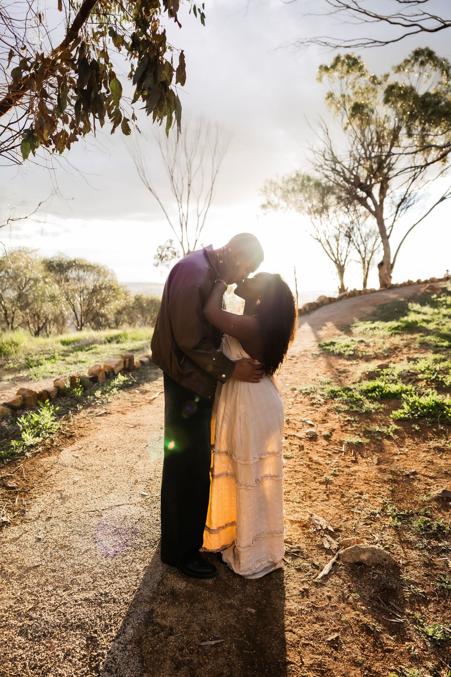 A couple shares a romantic kiss backlit by golden sunlight on a scenic outdoor dirt path surrounded by trees.