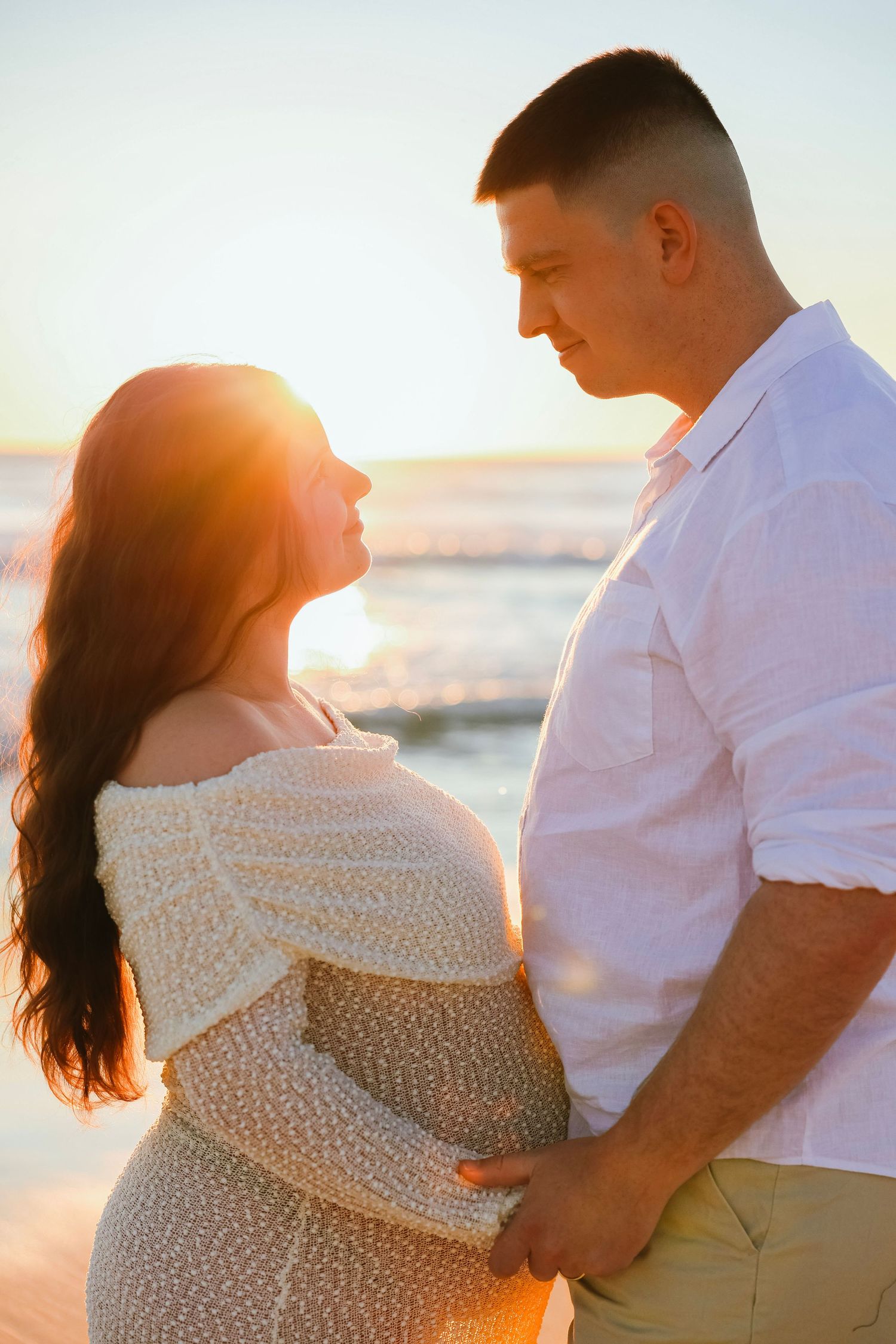 Pregnant couple sharing a tender moment at sunset on the beach, golden light glowing behind them.