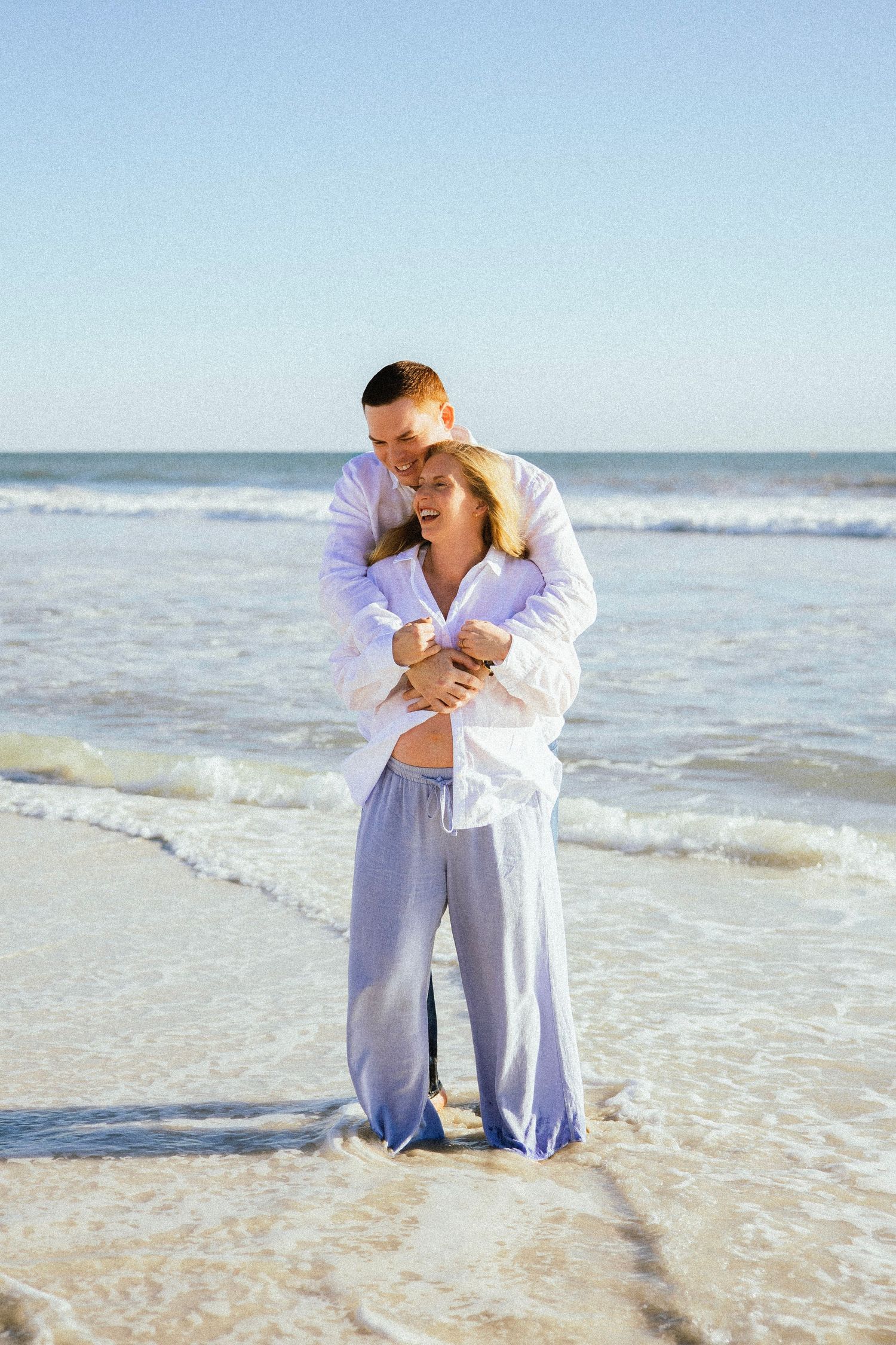 Couple embracing on a sunny beach, woman in white shirt and blue pants, waves gently washing ashore.