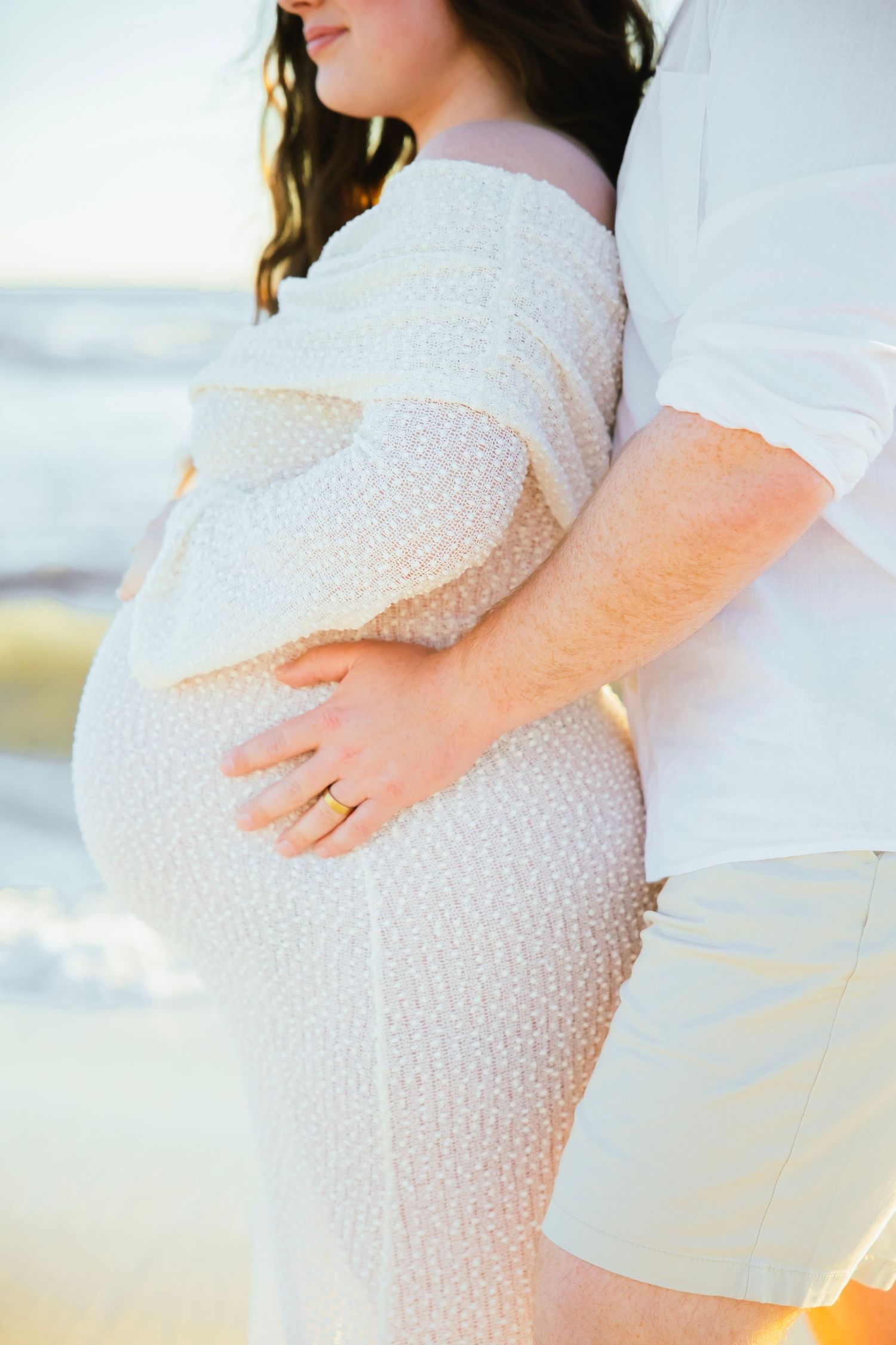 Pregnant woman in white textured dress embraced by partner on beach, golden sunset light, maternity photo session.