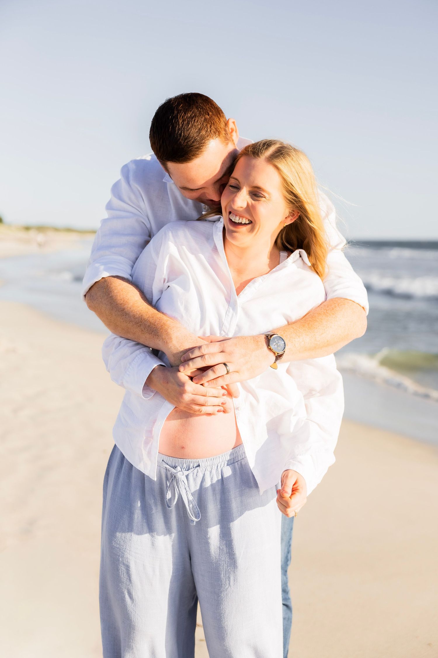 Man hugging pregnant woman from behind on beach, both smiling joyfully in white shirts by the ocean.