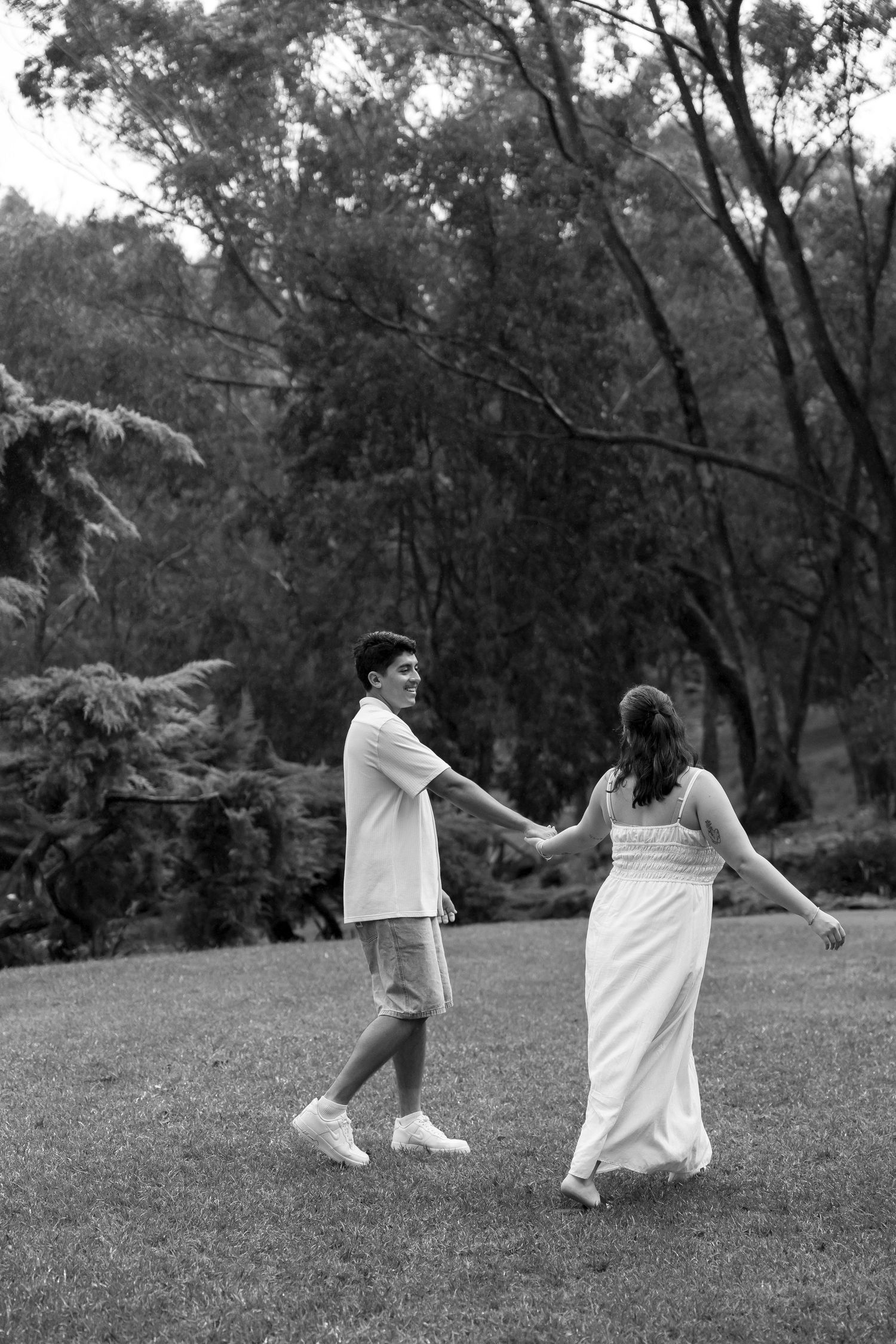 Black and white photo of a couple holding hands and walking through a lush green park.
