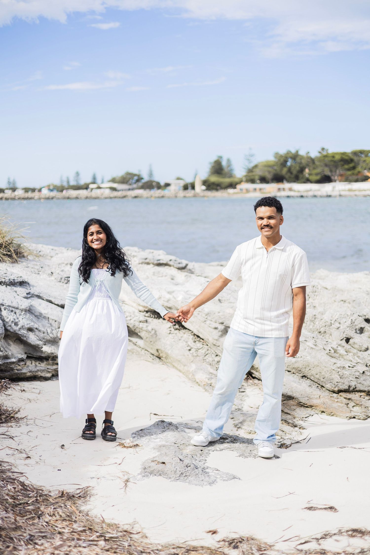 A couple holds hands while walking along a rocky shoreline with calm blue water behind them.