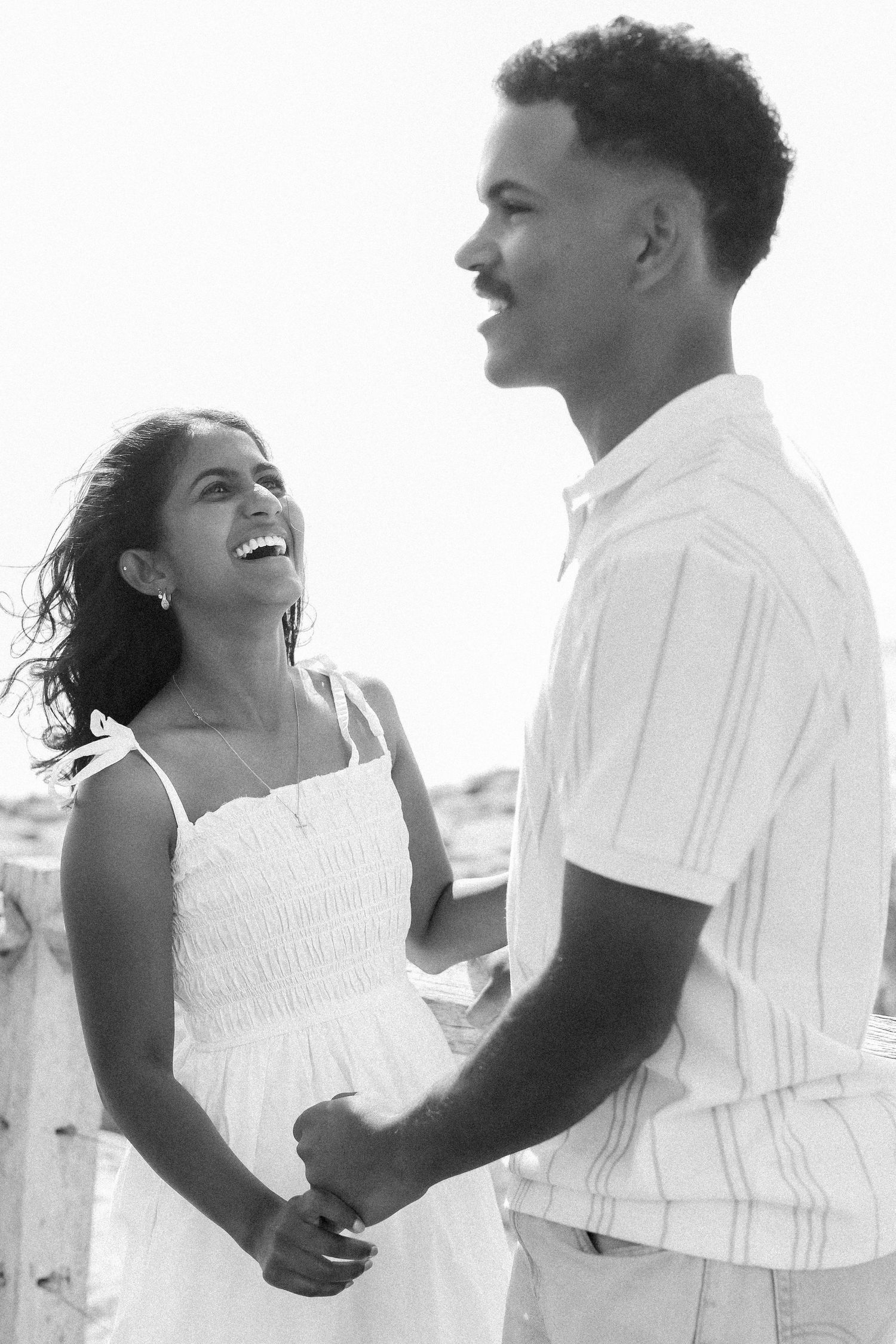 Black and white photo of a laughing couple holding hands at the beach in white outfits.